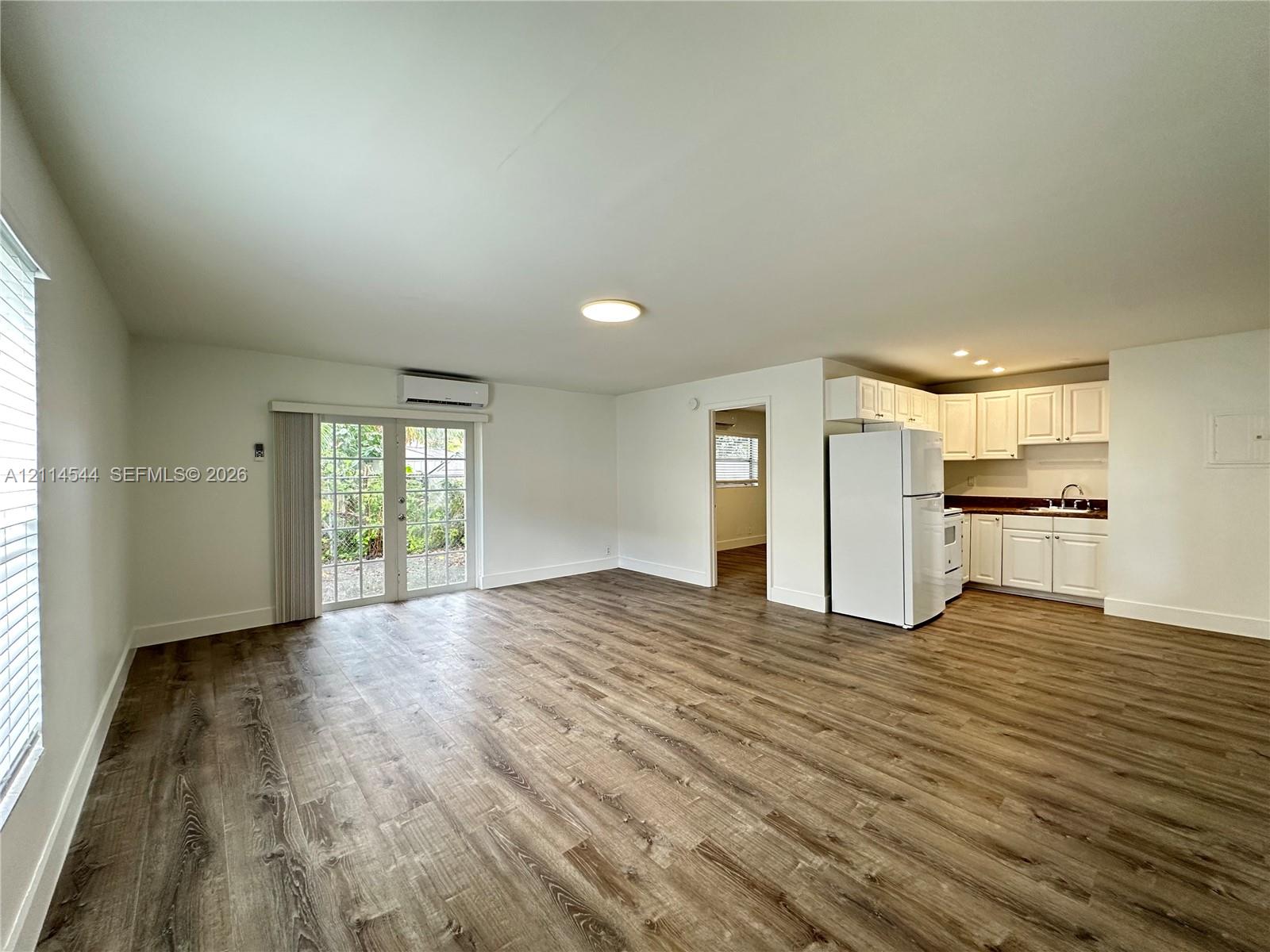 Open-concept interior featuring wood-finish flooring, glass-paned double doors, wall-mounted climate control unit, and white cabinetry with integrated kitchen appliances Open-concept interior featuring wood-finish flooring, glass-paned double doors, wall-mounted climate control unit, and white cabinetry with integrated kitchen appliances