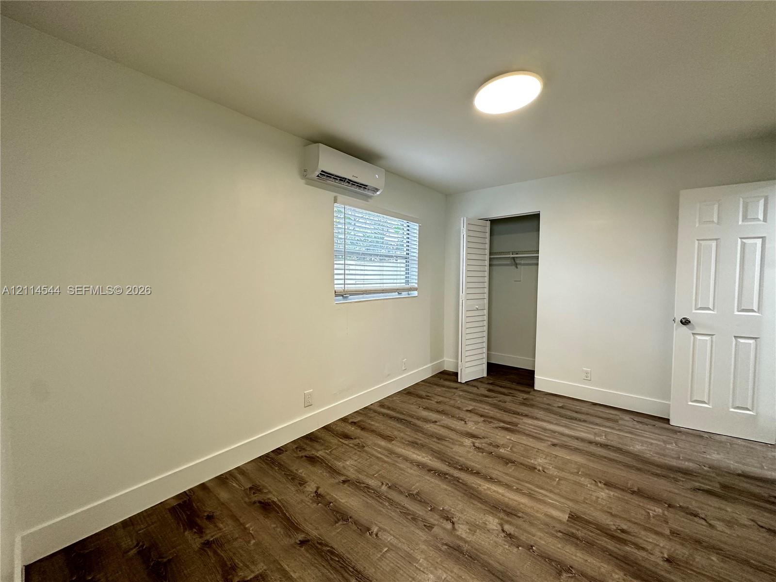 Room featuring wood-finish flooring, a wall-mounted ductless air conditioning unit, flush-mount ceiling lighting, a louvered-door closet, and a six-panel interior door Room featuring wood-finish flooring, a wall-mounted ductless air conditioning unit, flush-mount ceiling lighting, a louvered-door closet, and a six-panel interior door