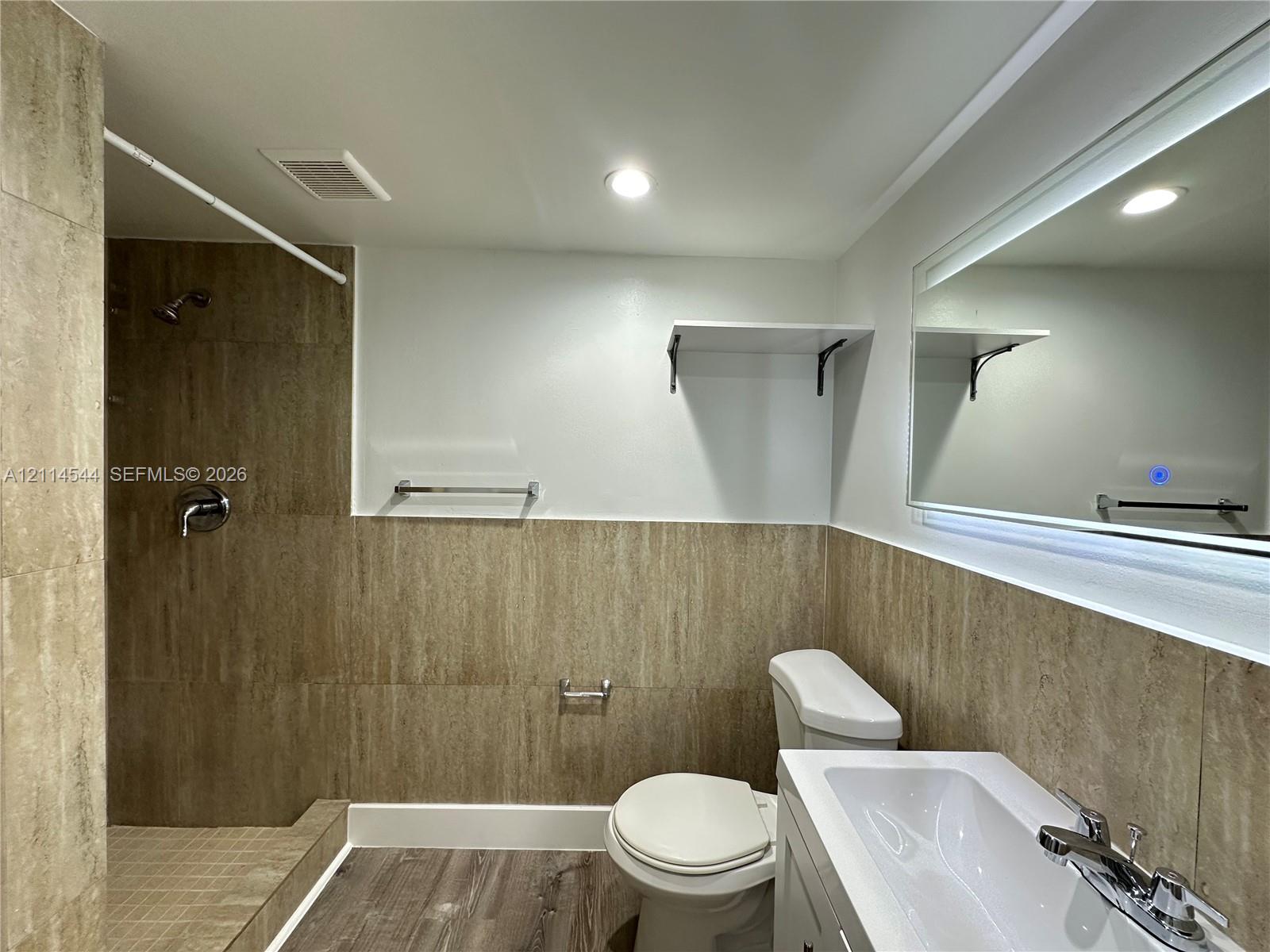 Bathroom featuring walk-in shower with floor-to-ceiling textured tile, wood-finish flooring, white vanity with integrated sink, and an illuminated wall-mounted mirror