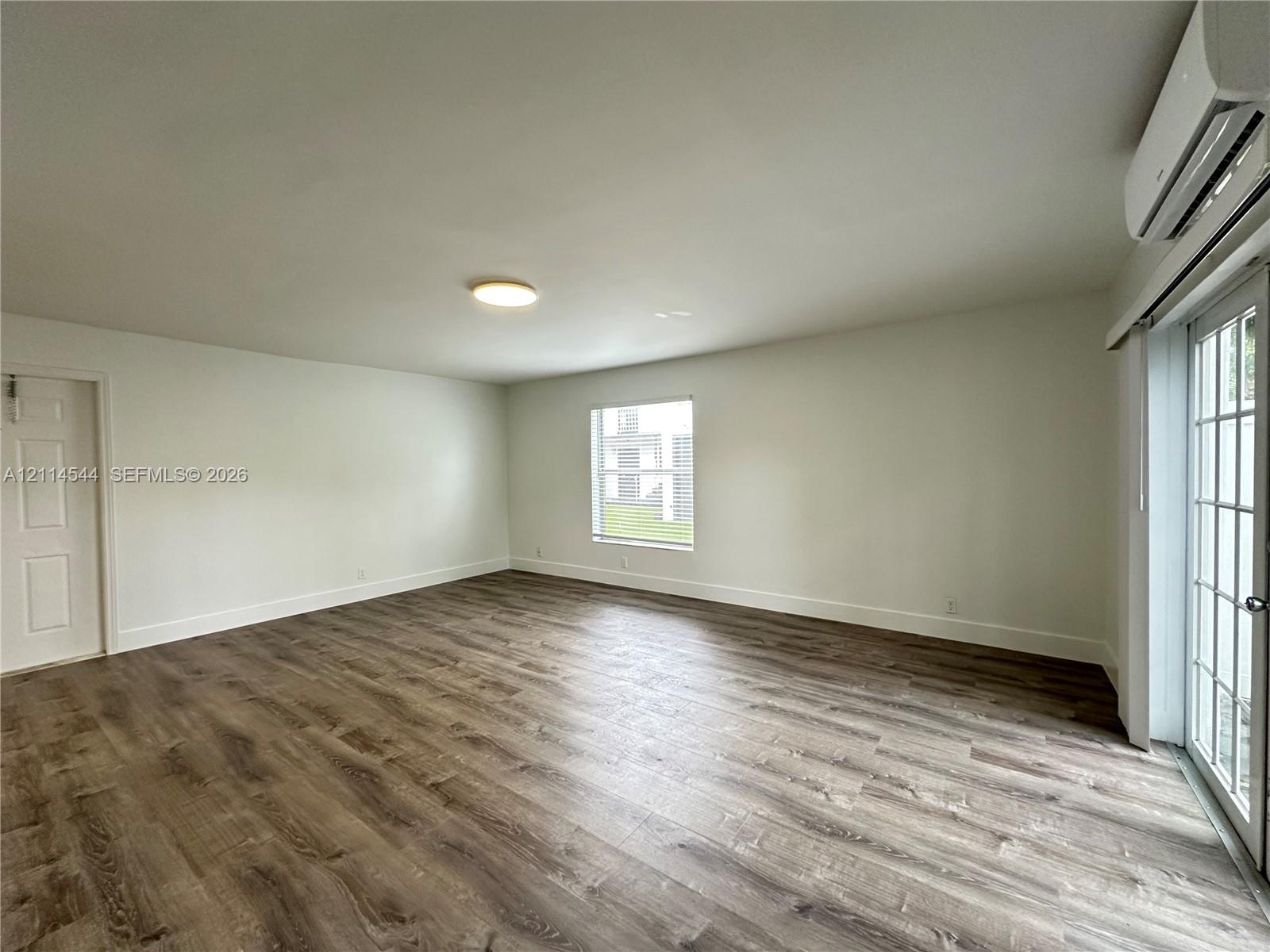 Neutral-toned interior featuring wood-finish flooring, a wall-mounted ductless heating and cooling unit, flush-mount ceiling lighting, and a glass-paned exterior door Neutral-toned interior featuring wood-finish flooring, a wall-mounted ductless heating and cooling unit, flush-mount ceiling lighting, and a glass-paned exterior door