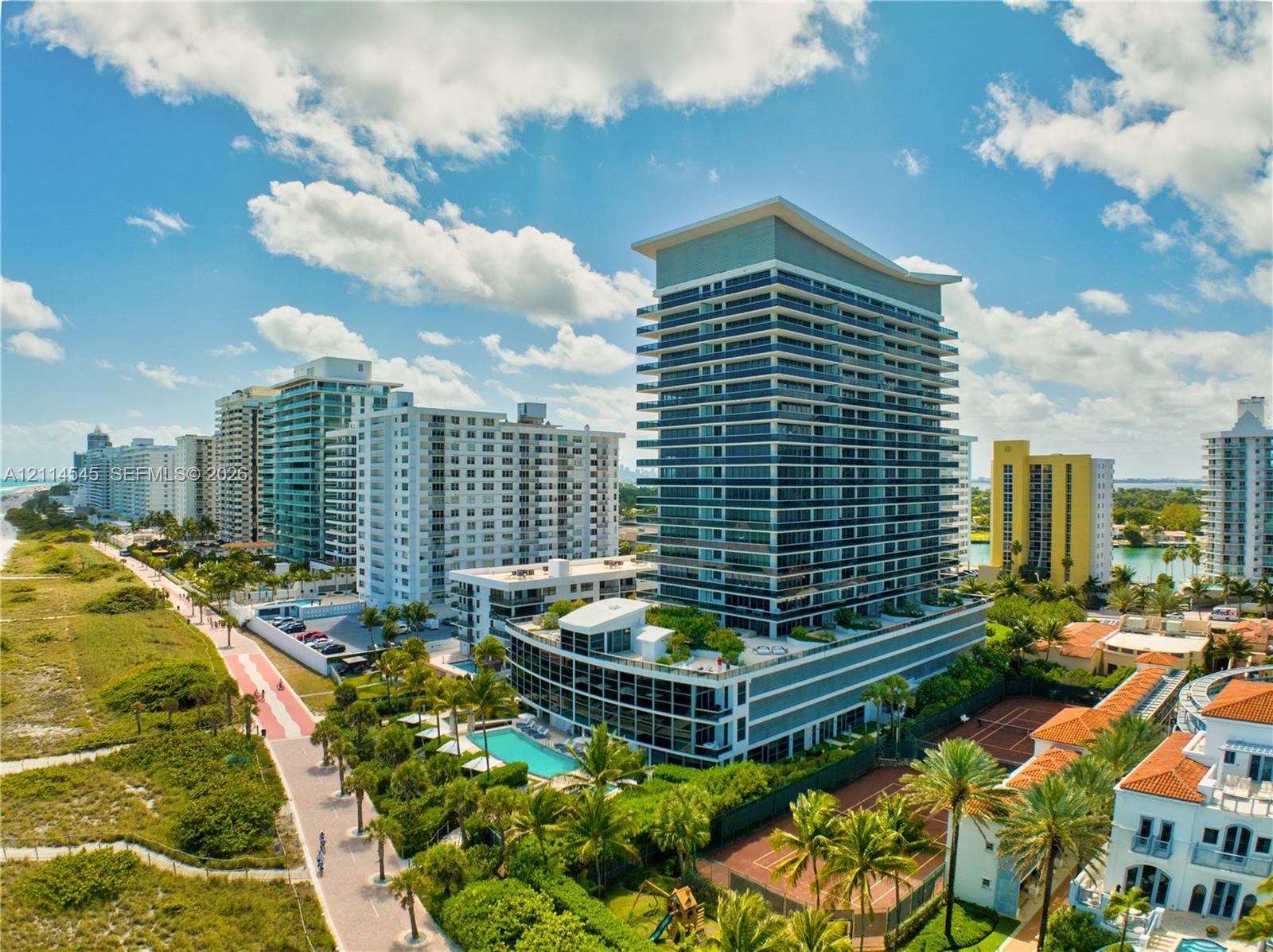 High-rise residential building featuring expansive glass balconies, an outdoor swimming pool, tennis courts, and landscaped grounds situated on a coastal site