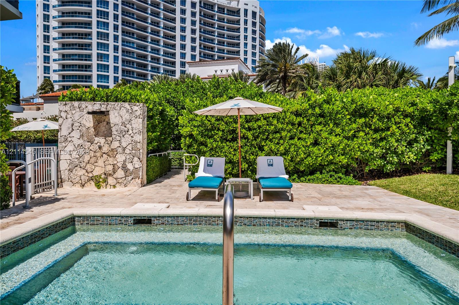 Outdoor spa with a tile-trimmed edge, natural stone patio, privacy hedge, and integrated stone water feature
