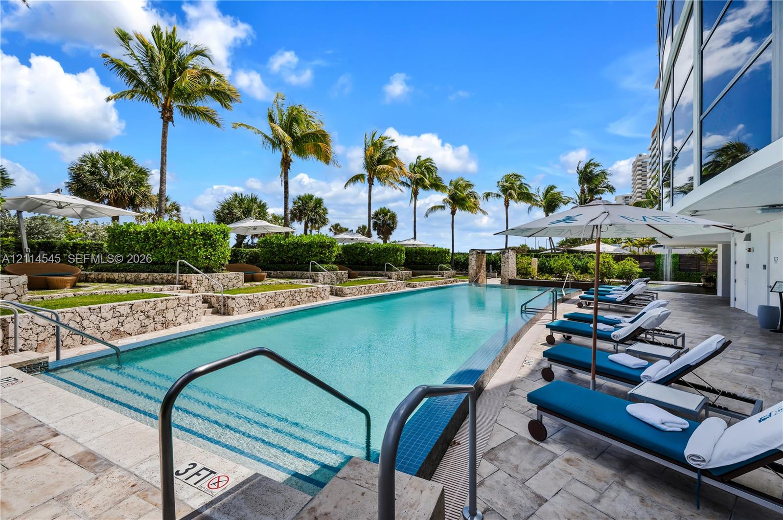 Rectangular swimming pool featuring a tiled waterline, stone-finished decking, tiered landscape planters, and palm tree-lined surroundings