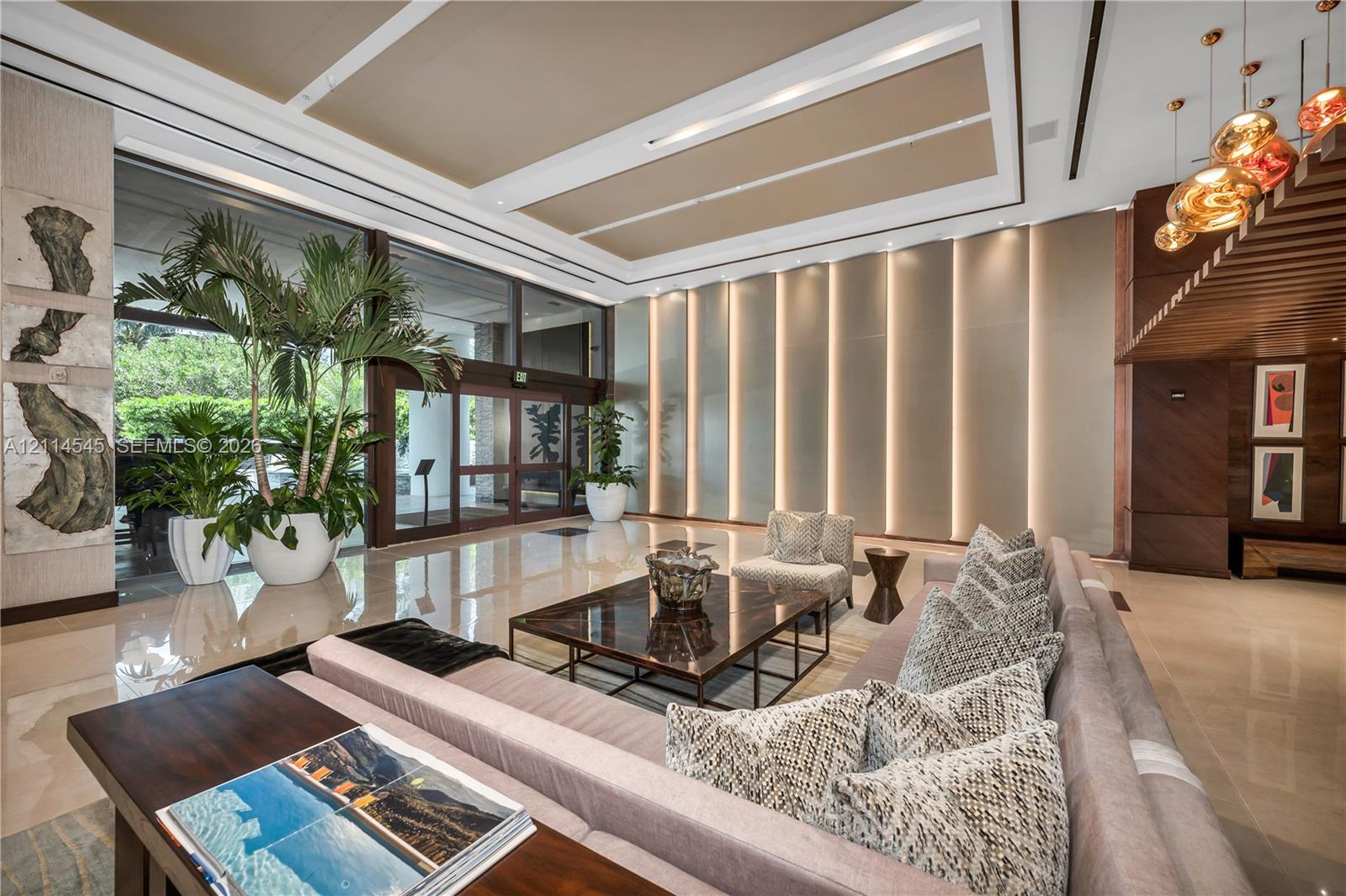 High-ceilinged foyer featuring polished stone flooring, recessed coffered ceiling, wall-integrated vertical lighting, floor-to-ceiling glass entry doors, and wood-slat ceiling accents