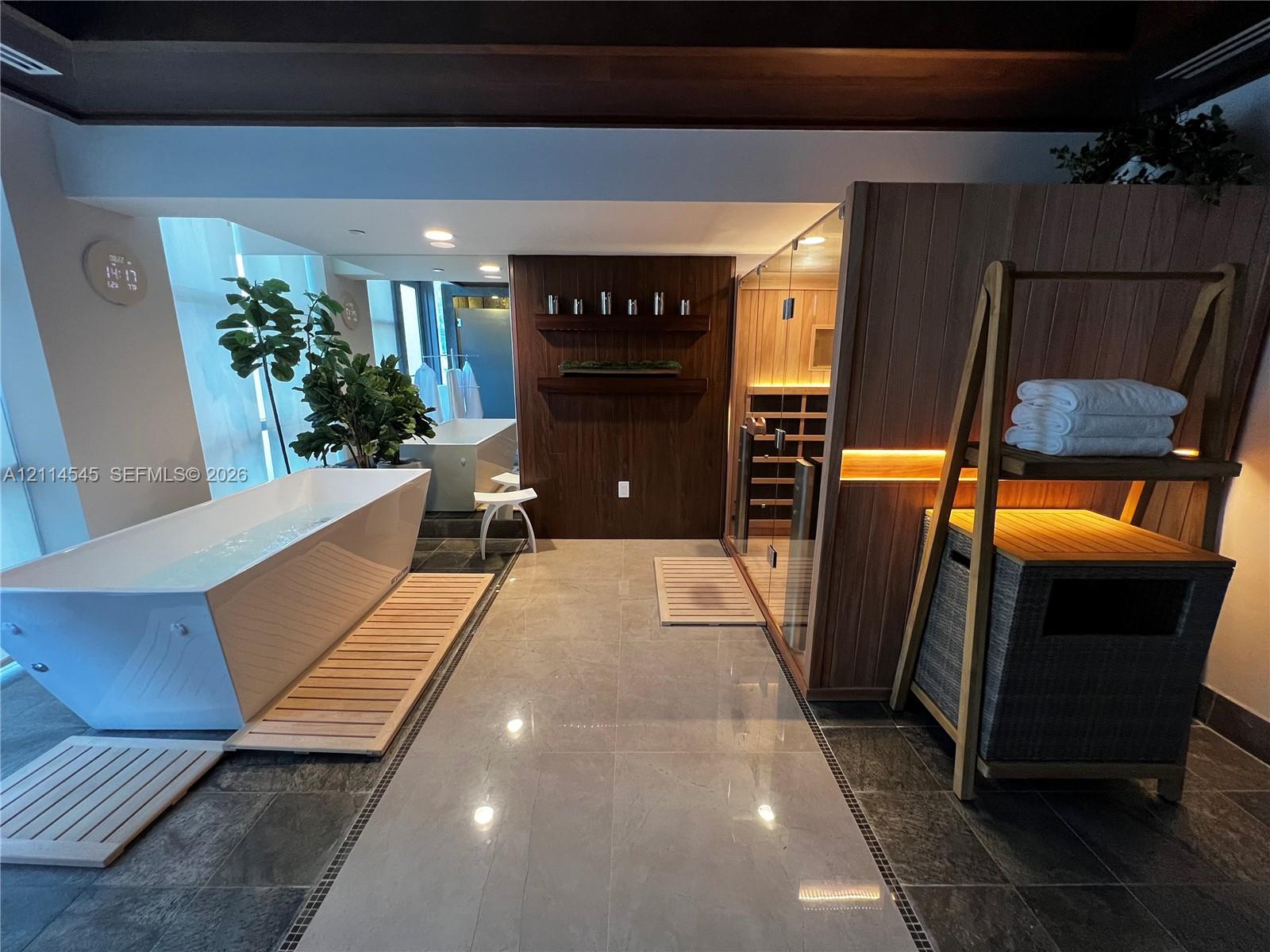 Freestanding soaking tub, glass-enclosed wood sauna with interior lighting, wood-paneled accent wall with built-in shelving, large-format tile flooring, and dark wood ceiling beams