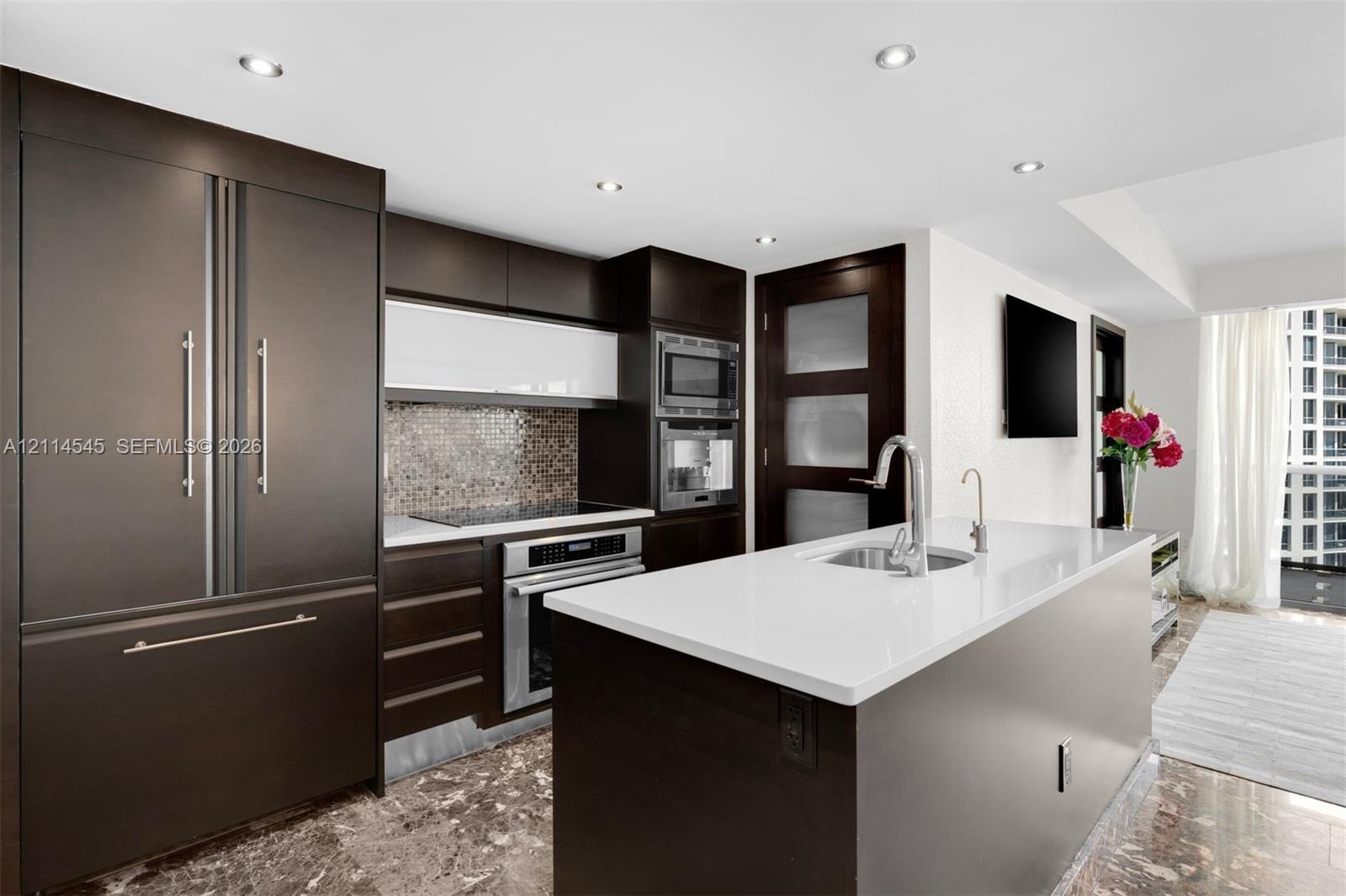 Modern kitchen featuring dark cabinetry, quartz-topped center island with integrated sink, stainless steel built-in appliances, recessed lighting, and stone flooring