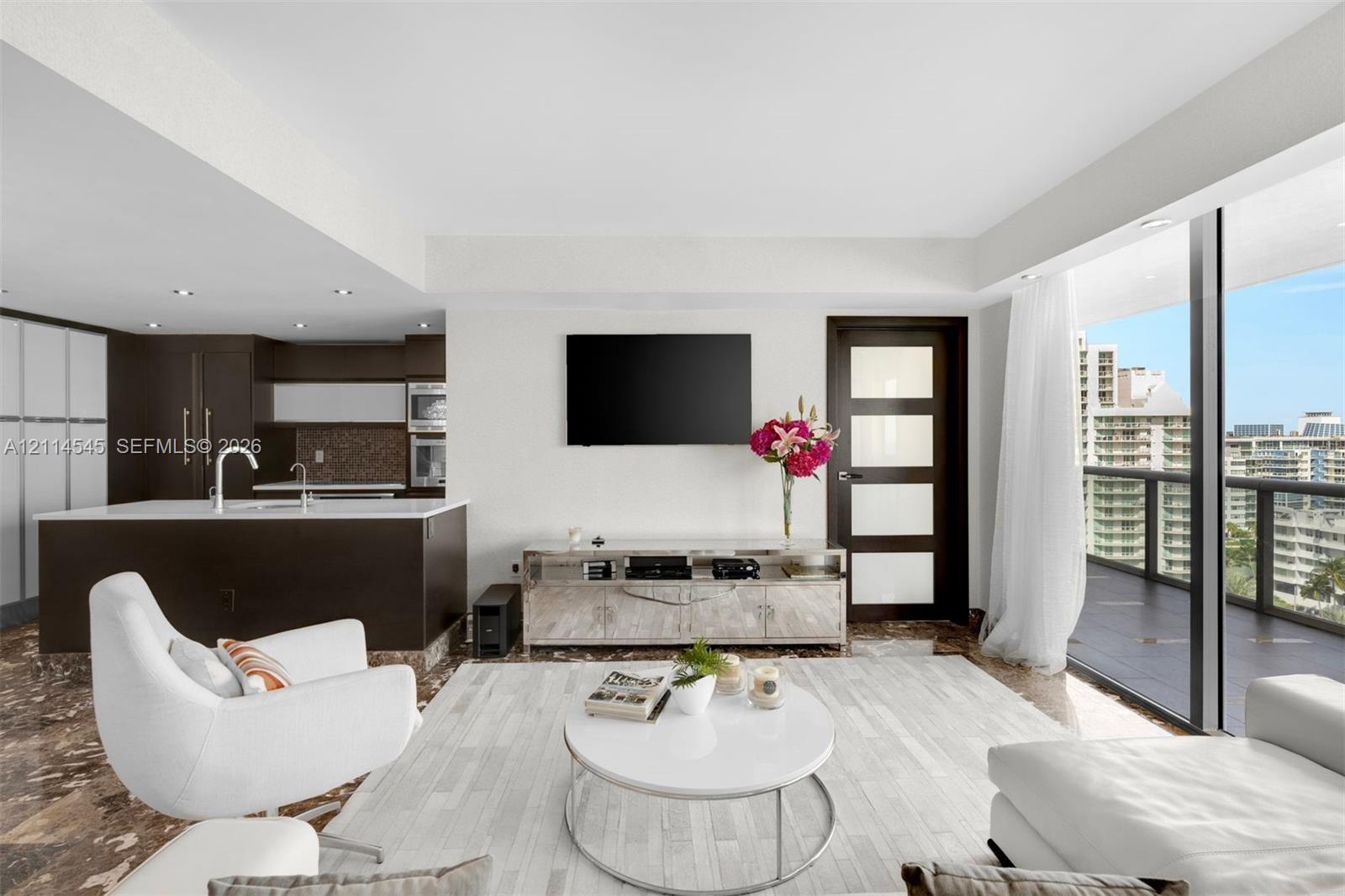 Open-concept living space featuring stone flooring, a kitchen island with integrated sink, recessed ceiling lighting, a five-panel frosted glass door, and expansive floor-to-ceiling windows leading to a covered balcony