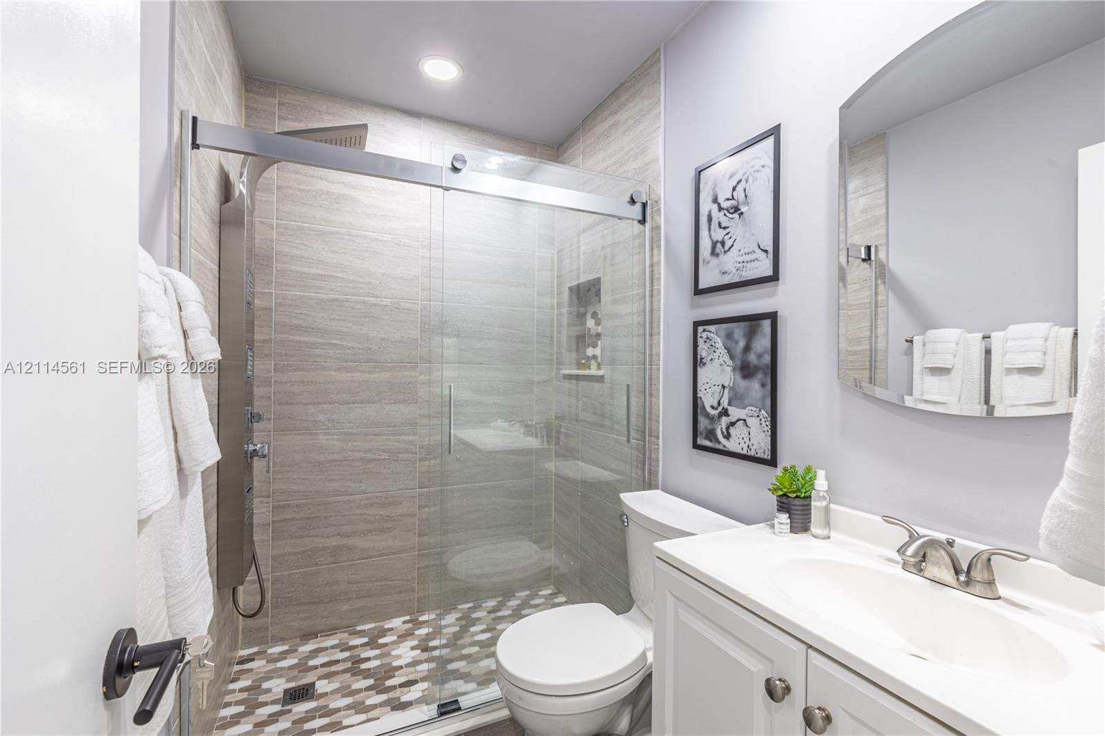 Bathroom featuring a glass-enclosed shower with horizontal wall tile, mosaic floor tiling, built-in recessed shelving, a multi-function shower panel, and a vanity with a white countertop