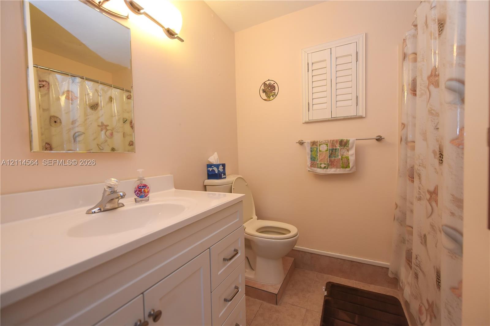 Bathroom featuring a white vanity with an integrated basin, wall-mounted mirror, tiered light fixture, shutters, and ceramic tile flooring