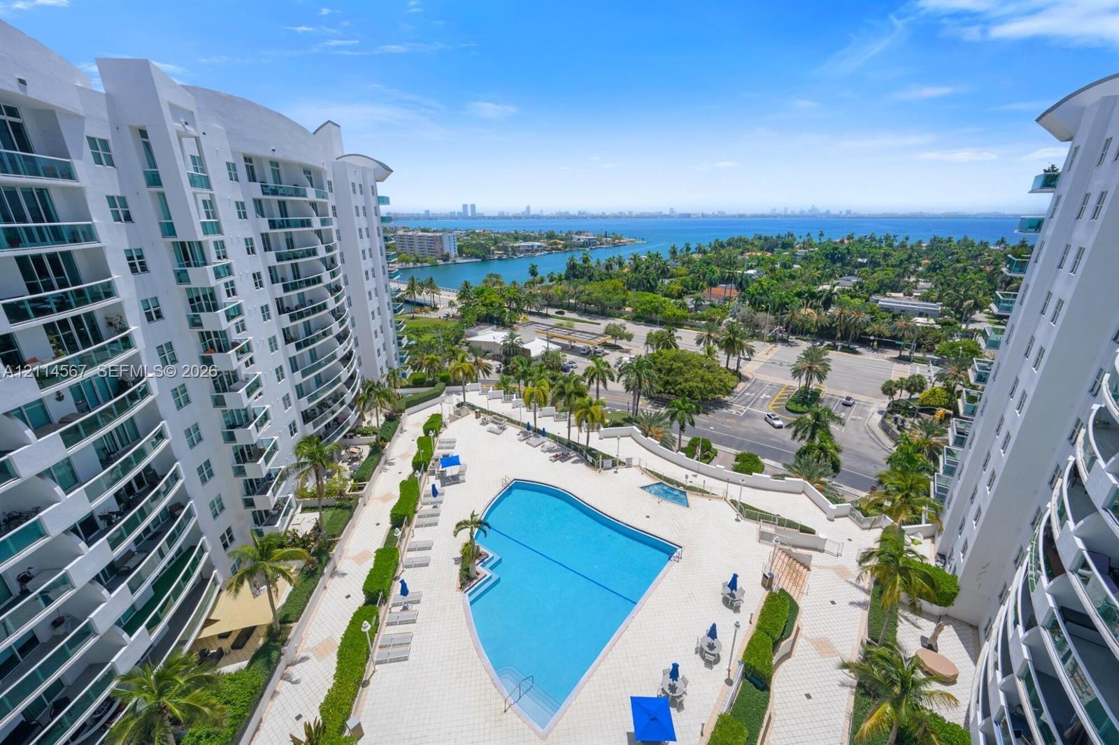 Outdoor swimming pool, paved sundeck, mature palm trees, multi-story building architecture with balconies, and coastal water backdrop