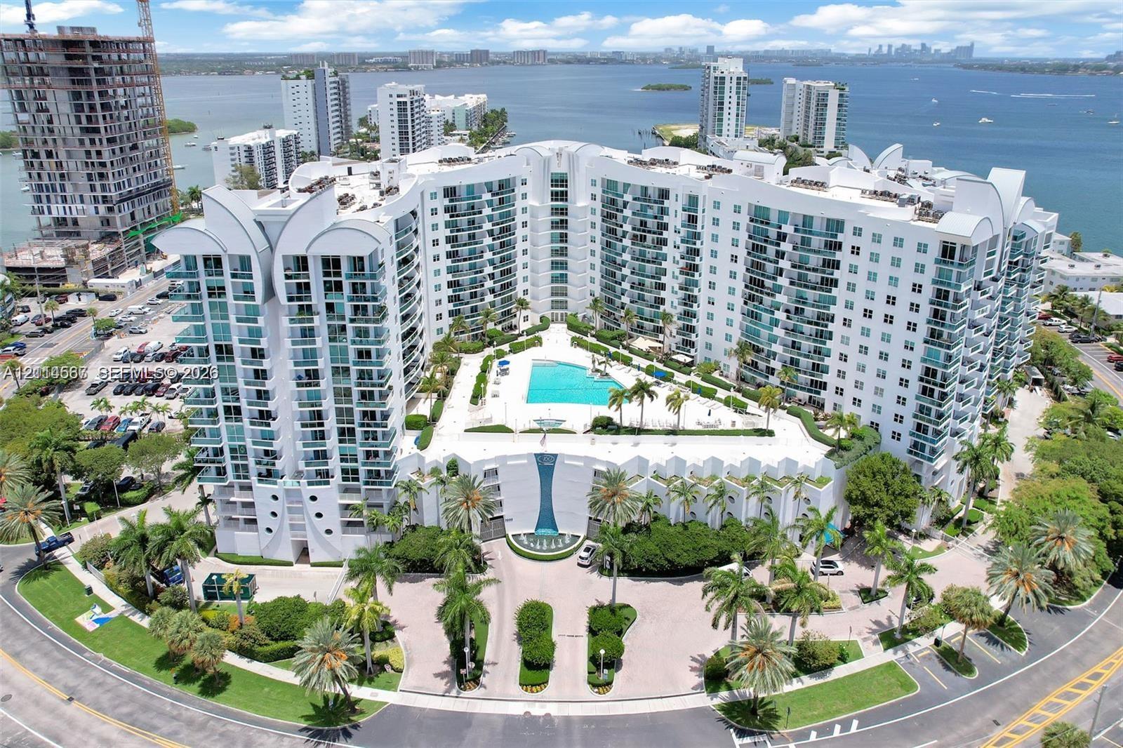 Multi-story waterfront condominium building featuring an expansive outdoor swimming pool, paved motor court, manicured palm landscaping, and wrap-around private balconies