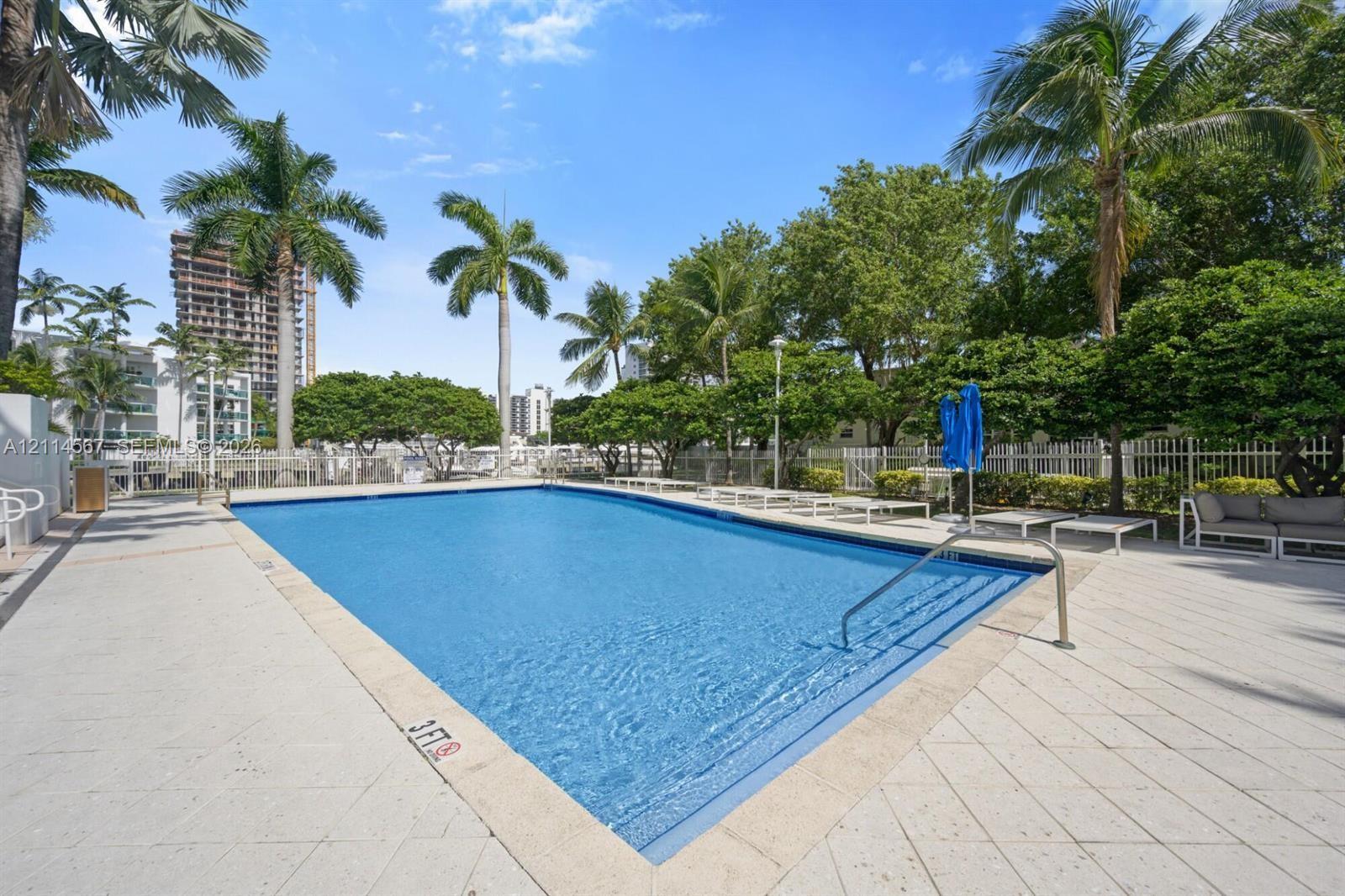 Rectangular swimming pool featuring a tiled deck, metal handrail, palm tree landscaping, and perimeter fencing