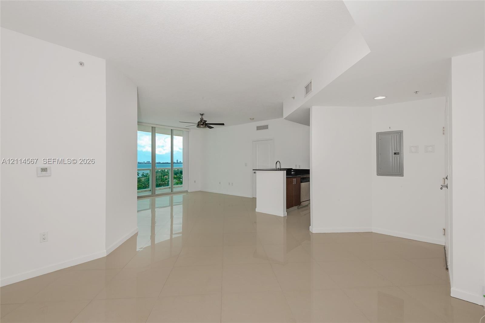 Open-concept interior featuring large-format tile flooring, floor-to-ceiling glass windows, a ceiling fan, and a kitchen peninsula with dark countertops and wood-finish cabinetry