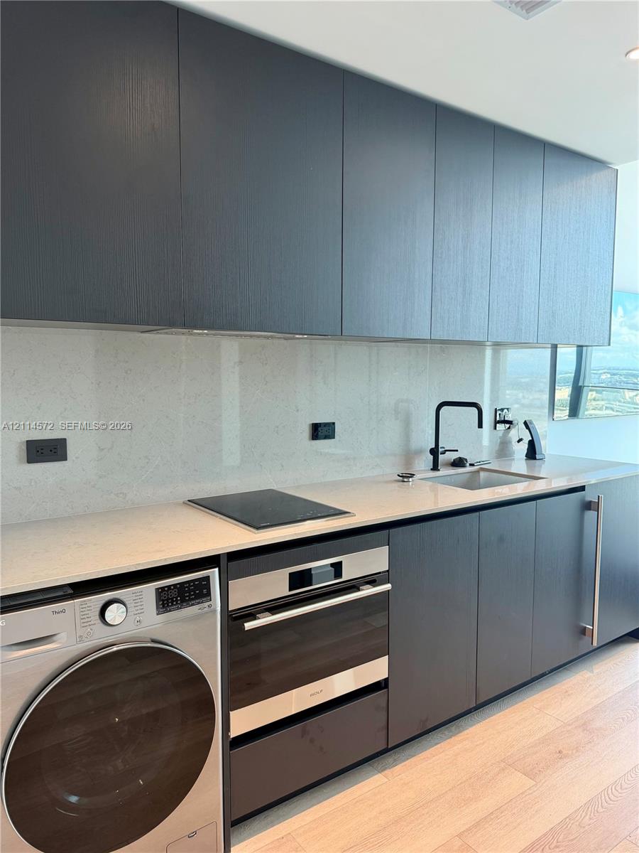 Contemporary kitchen featuring charcoal-toned flat-panel cabinetry, light-toned stone-surface countertops, integrated cooktop, stainless steel wall oven, and light wood-finish flooring