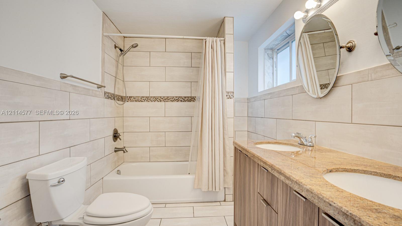 Dual-sink vanity with stone countertop, large-format tile surround, bathtub and shower combination, and oval wall-mounted mirrors