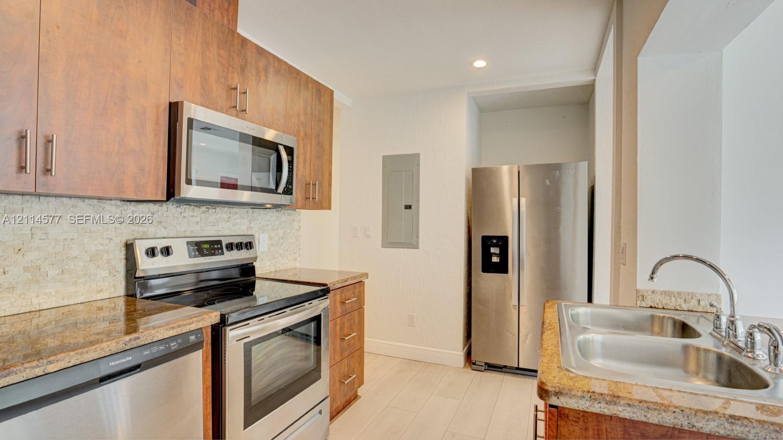 Kitchen featuring wood-finish cabinetry, stone-texture backsplash, stainless steel appliances, and solid-surface countertops with a dual-basin undermount sink