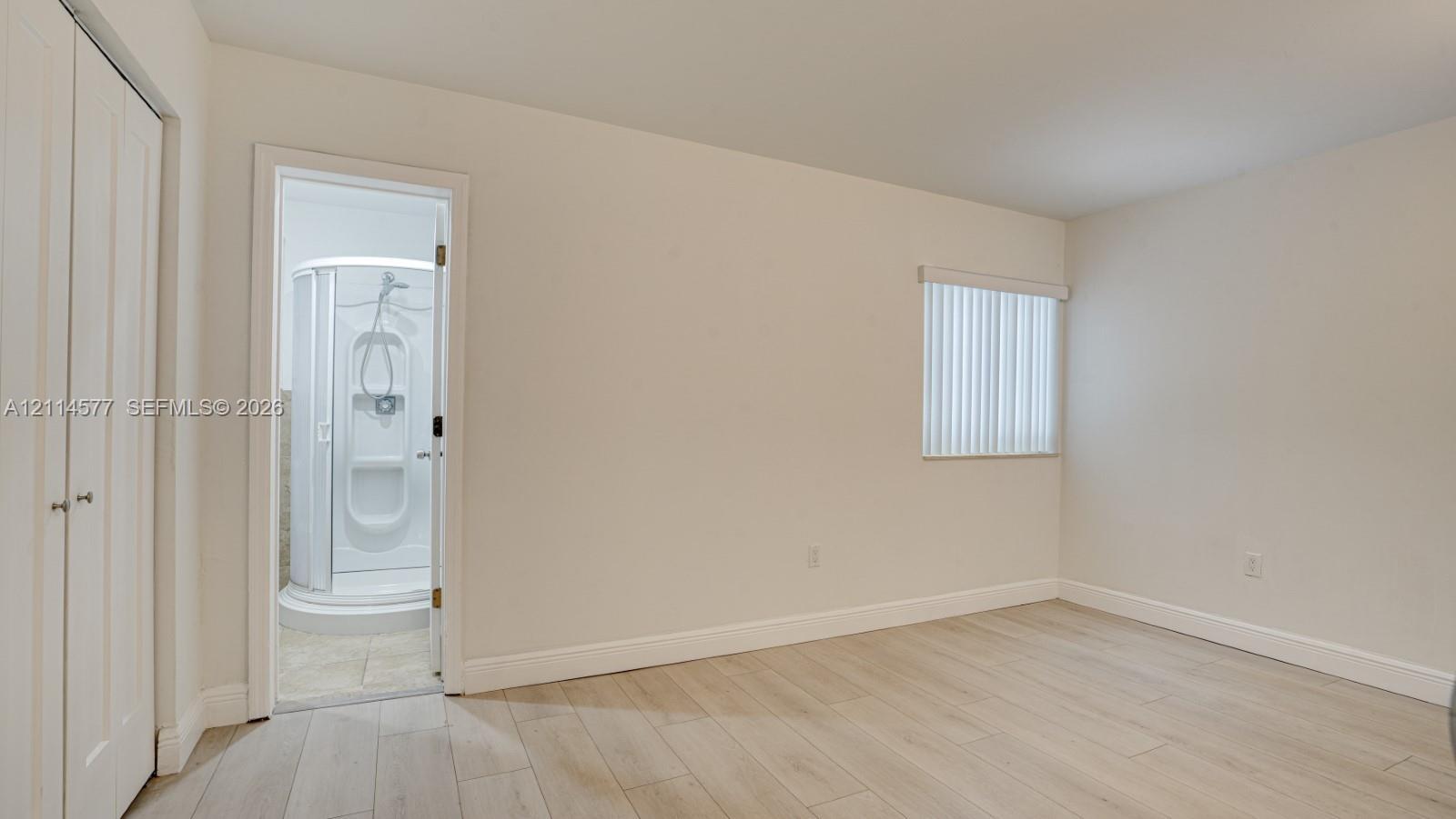 Room featuring wood-finish flooring, white double-door closet, and window with vertical blinds, accompanied by an enclosed shower stall with integrated shelving
