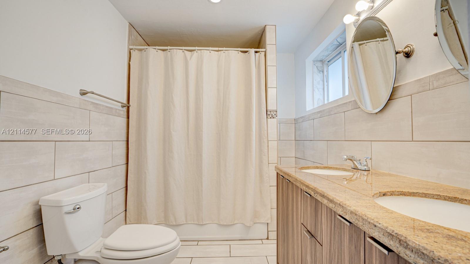 Dual-sink vanity with stone-surfaced countertop, wood-finish cabinetry, ceramic tile wall cladding, and walk-in shower enclosure