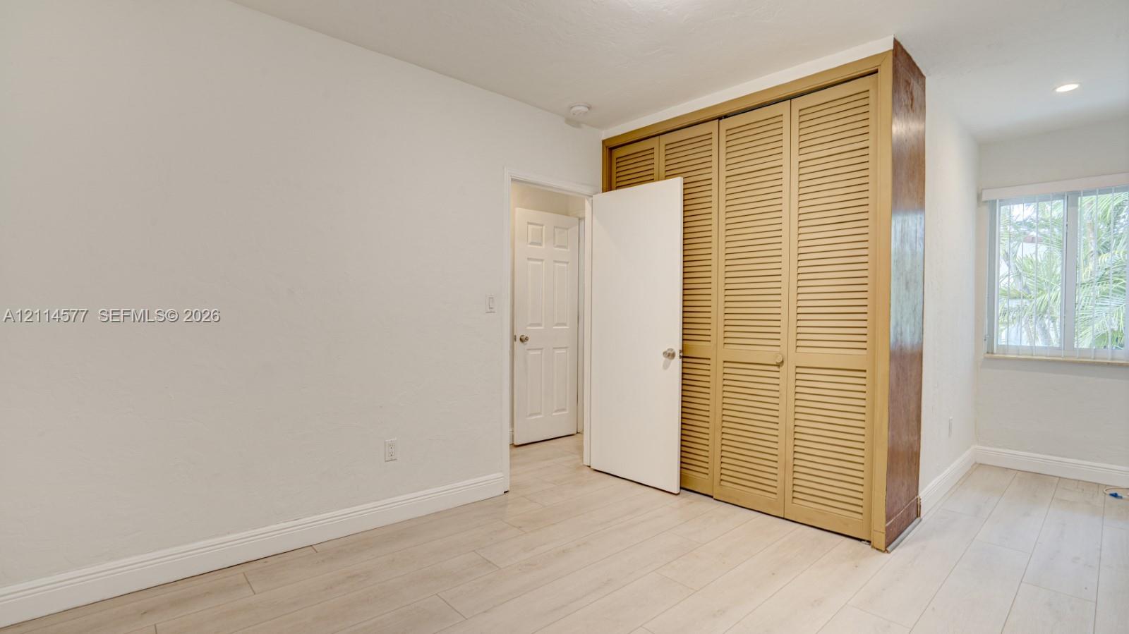 Neutral interior space featuring wood-finish flooring, louvered closet doors, and a window with white casing