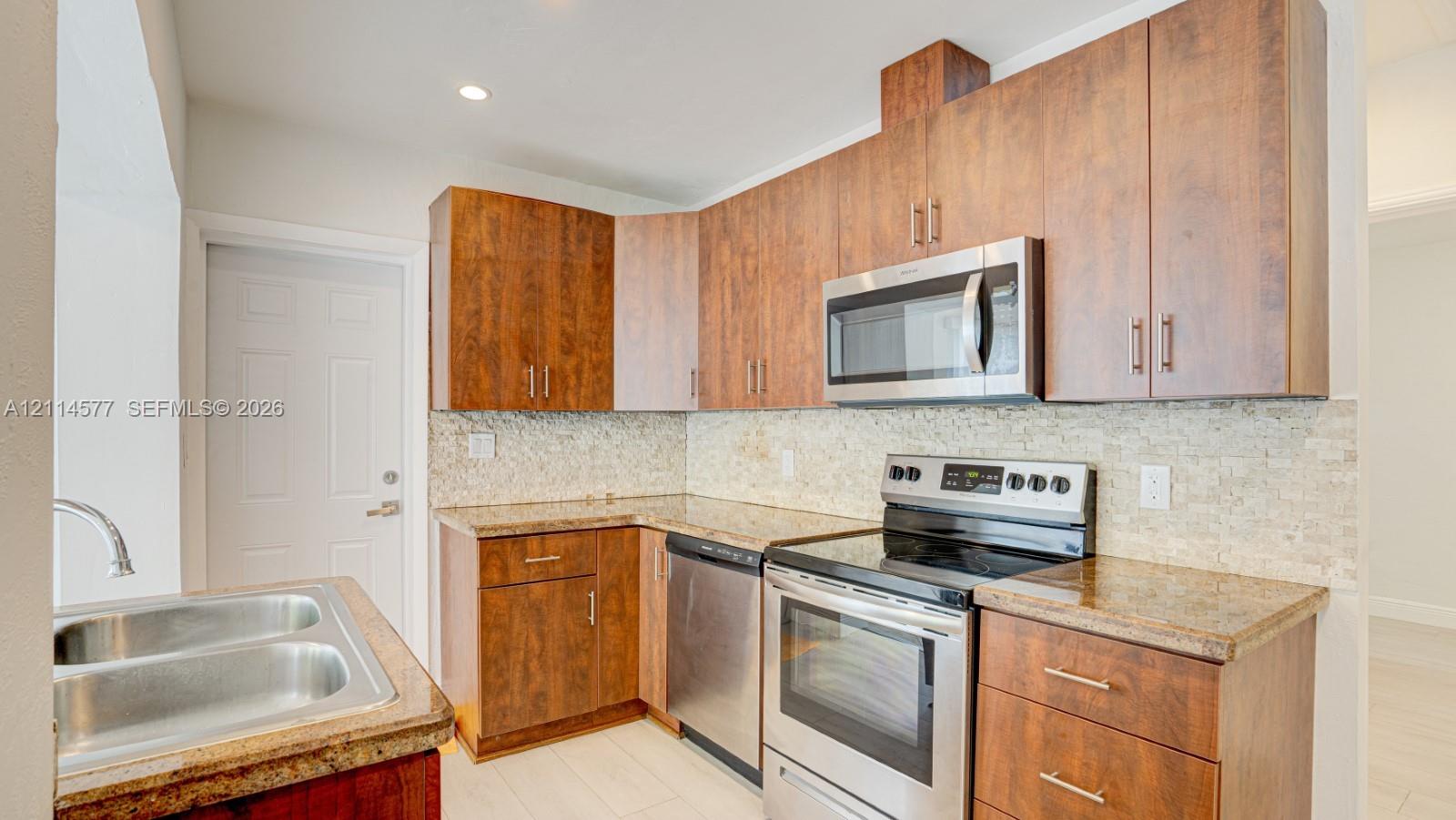 Kitchen featuring flat-panel wood-finish cabinetry, stone-surface countertops, textured stone backsplash, stainless steel appliances, and a double-basin sink