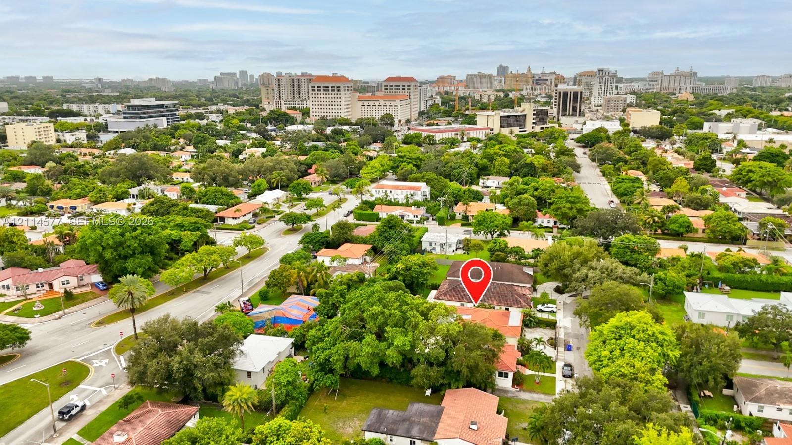 Aerial perspective of a residential property featuring a pitched roof, mature canopy trees, established landscaping, and paved street access