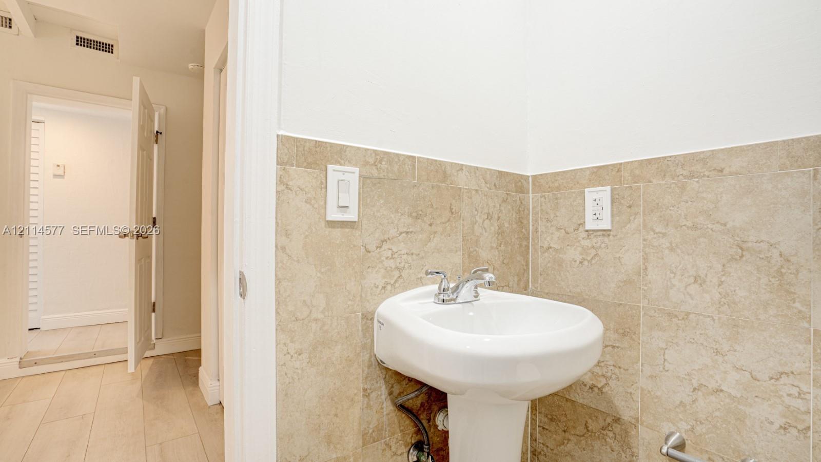 White pedestal sink featuring a dual-handle chrome faucet, complemented by neutral-toned tile wainscoting and light wood-finish flooring