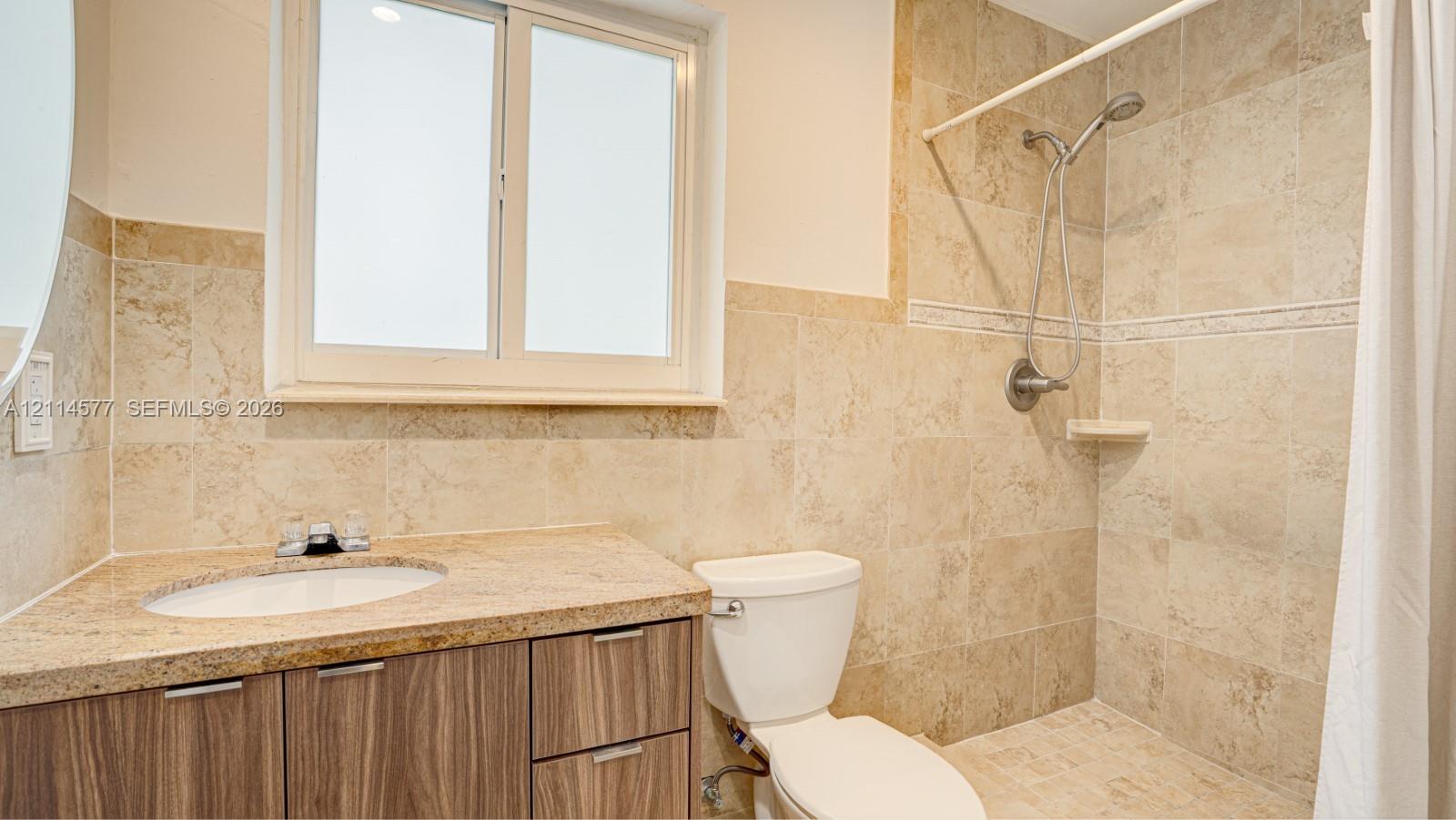 Bathroom featuring stone-tile wall surround, wood-finish vanity cabinetry, stone-slab countertop with integrated oval sink, and a handheld shower fixture