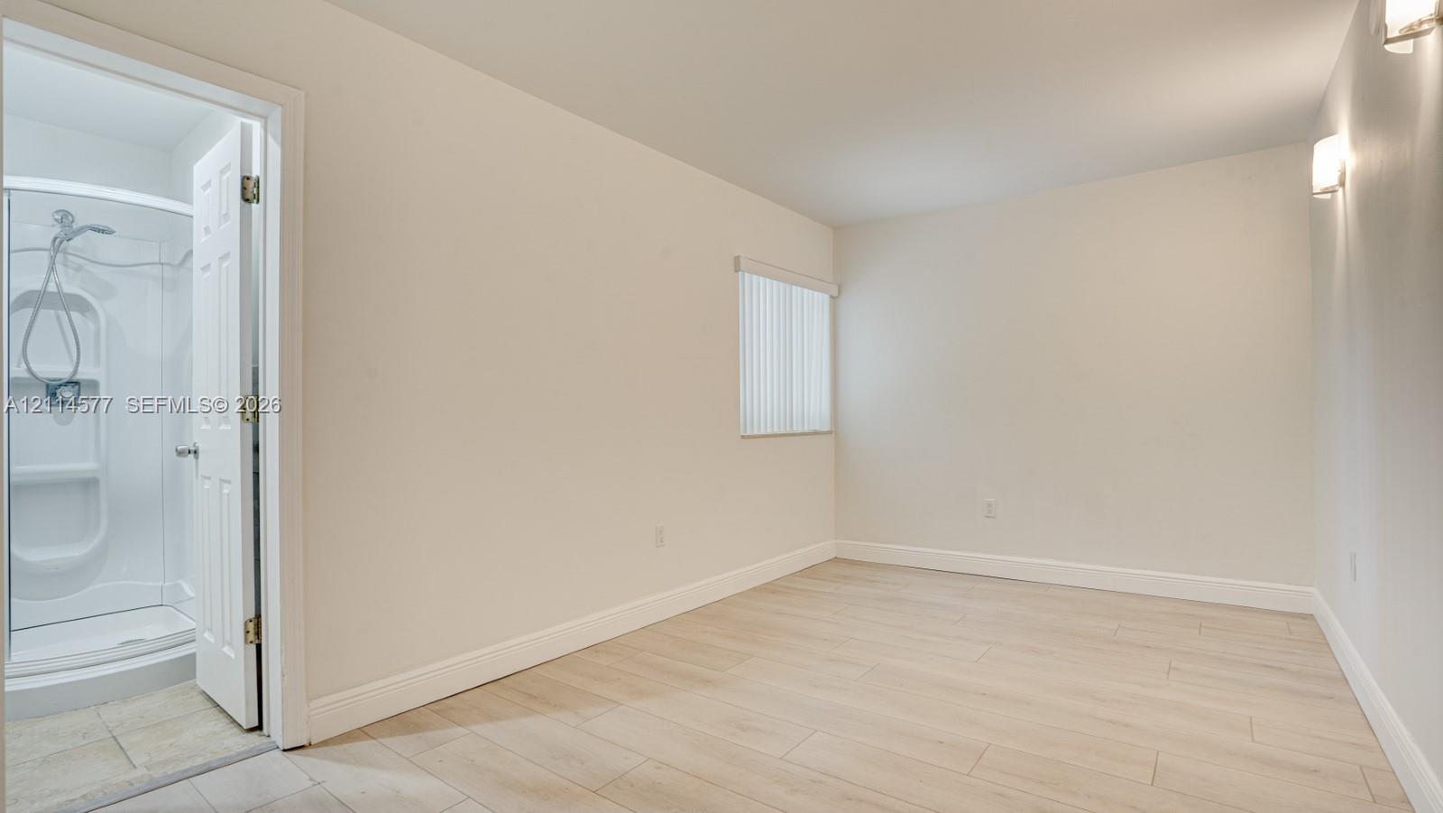 Neutral-toned room with wood-finish flooring, a vertical blind window covering, wall-mounted light fixtures, and a shower stall enclosure with a handheld faucet