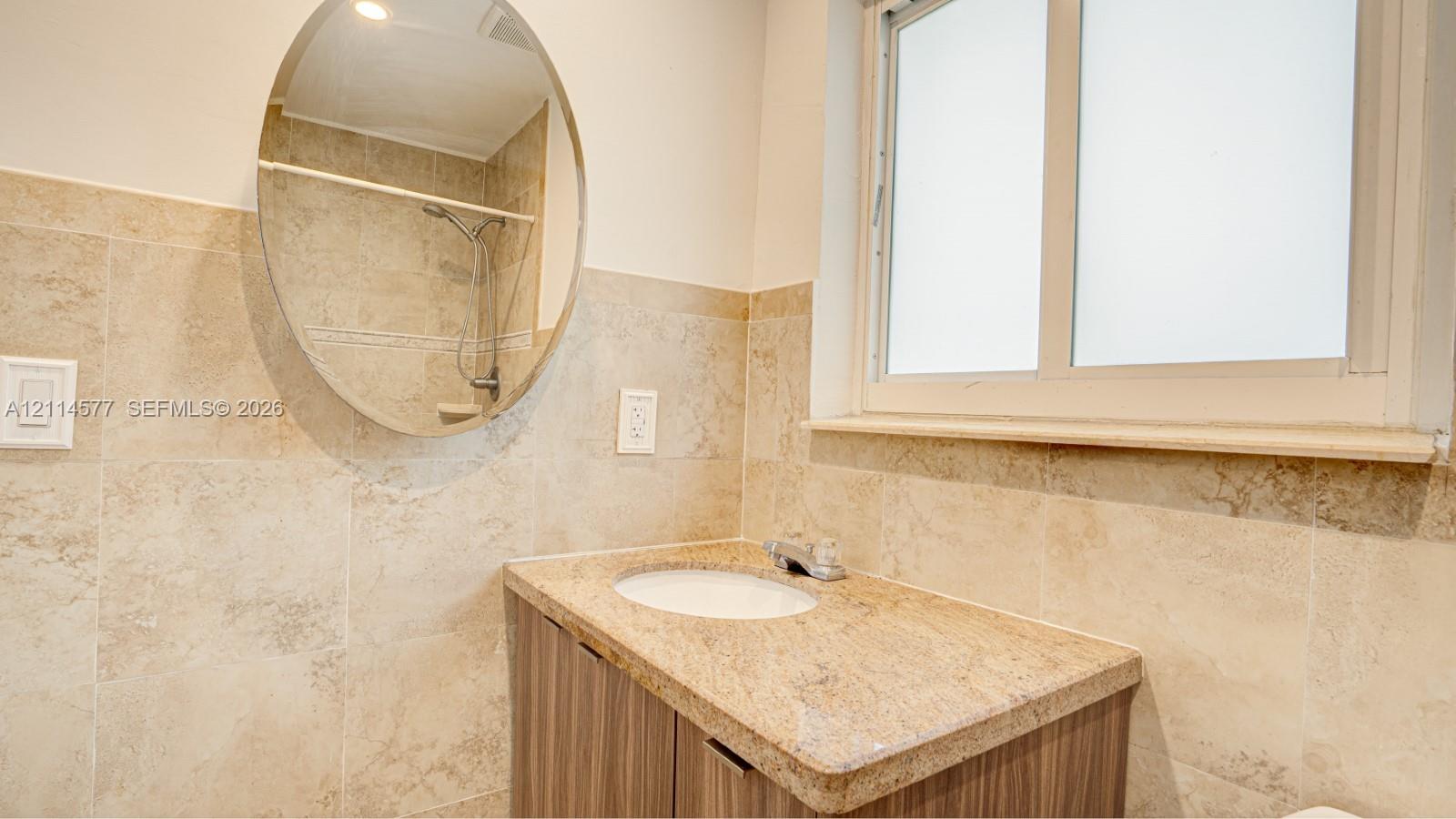 Stone-tiled walls, vanity with stone countertop and integrated sink, oval wall-mounted mirror, and frosted glass window
