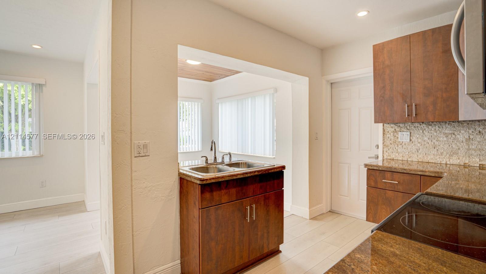 Kitchen featuring wood-finish cabinetry, stone-texture countertops, a dual-basin stainless steel sink, tiled backsplash, and light-toned wood-finish flooring
