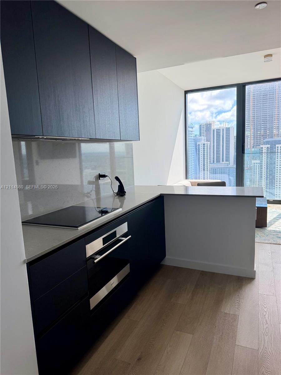 Kitchen featuring dark slab-style cabinetry, solid-surface countertops, integrated cooktop, stainless steel wall oven, and wood-finish flooring