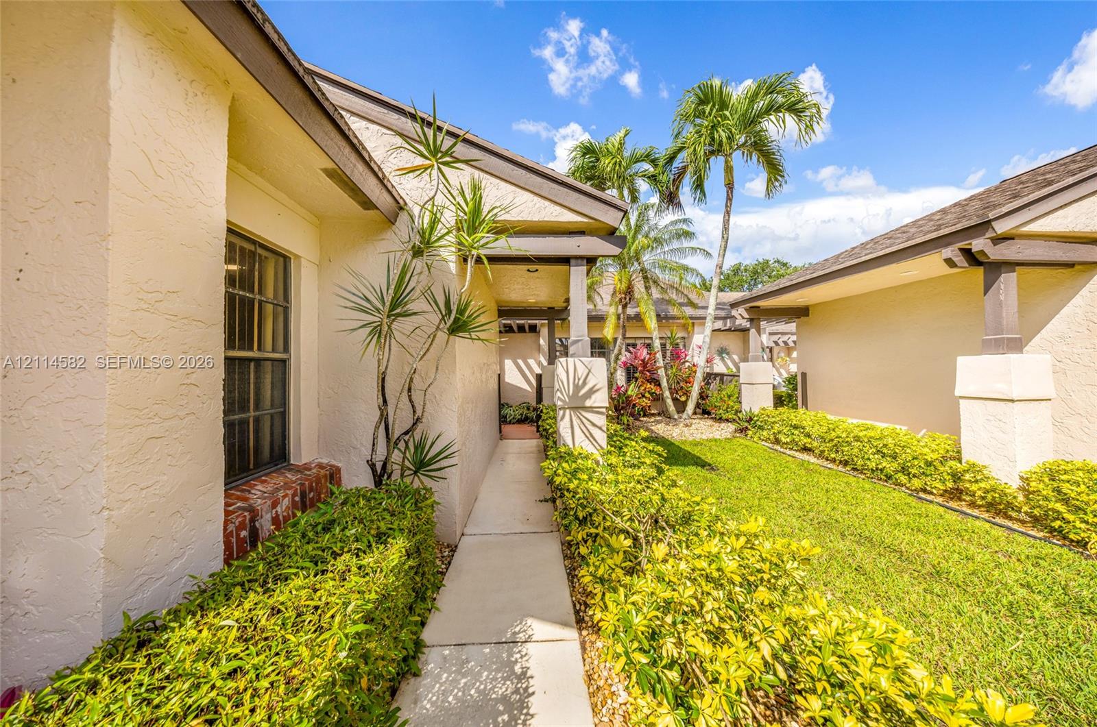 Stucco exterior with masonry accents, covered walkway, architectural support columns, and landscaped grounds featuring palm trees