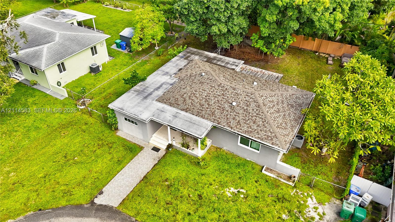 Aerial perspective of a single-story residence featuring a shingled roof, a paved walkway, a concrete-surfaced entryway, and an expansive yard with mature trees Aerial perspective of a single-story residence featuring a shingled roof, a paved walkway, a concrete-surfaced entryway, and an expansive yard with mature trees