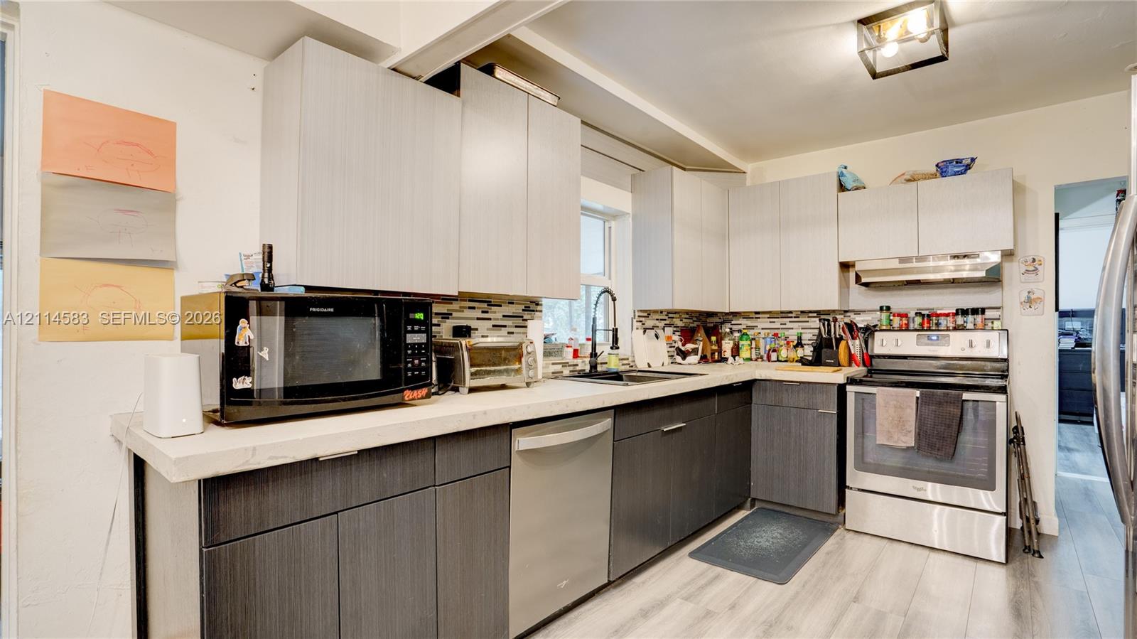 Kitchen featuring two-tone flat-panel cabinetry, light-toned stone-finish countertops, stainless steel appliances, multi-colored tile backsplash, and light-toned wood-finish flooring
