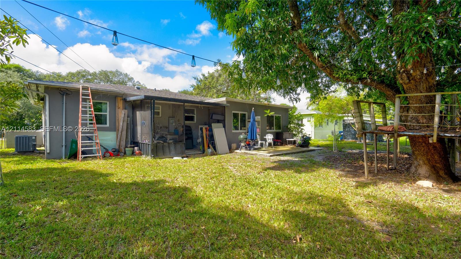Rear exterior featuring a screened-in patio, expansive grass lawn, mature shade trees, and an elevated wooden platform structure