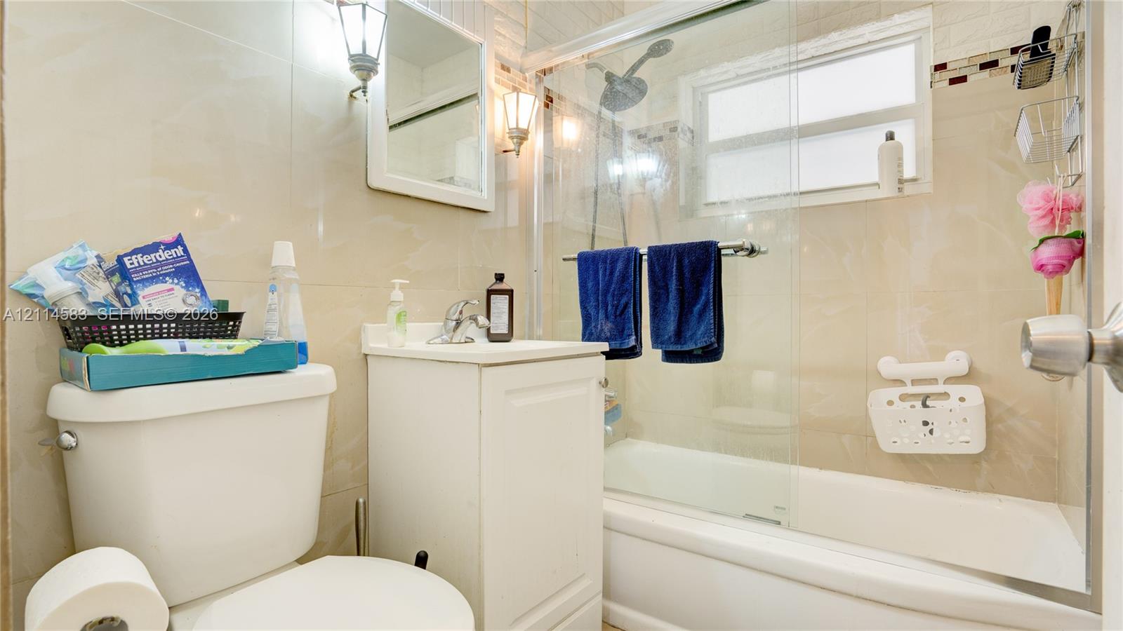 Bathroom featuring floor-to-ceiling neutral wall tile, white vanity cabinet with a chrome faucet, and a shower-tub combination enclosed by a glass panel door