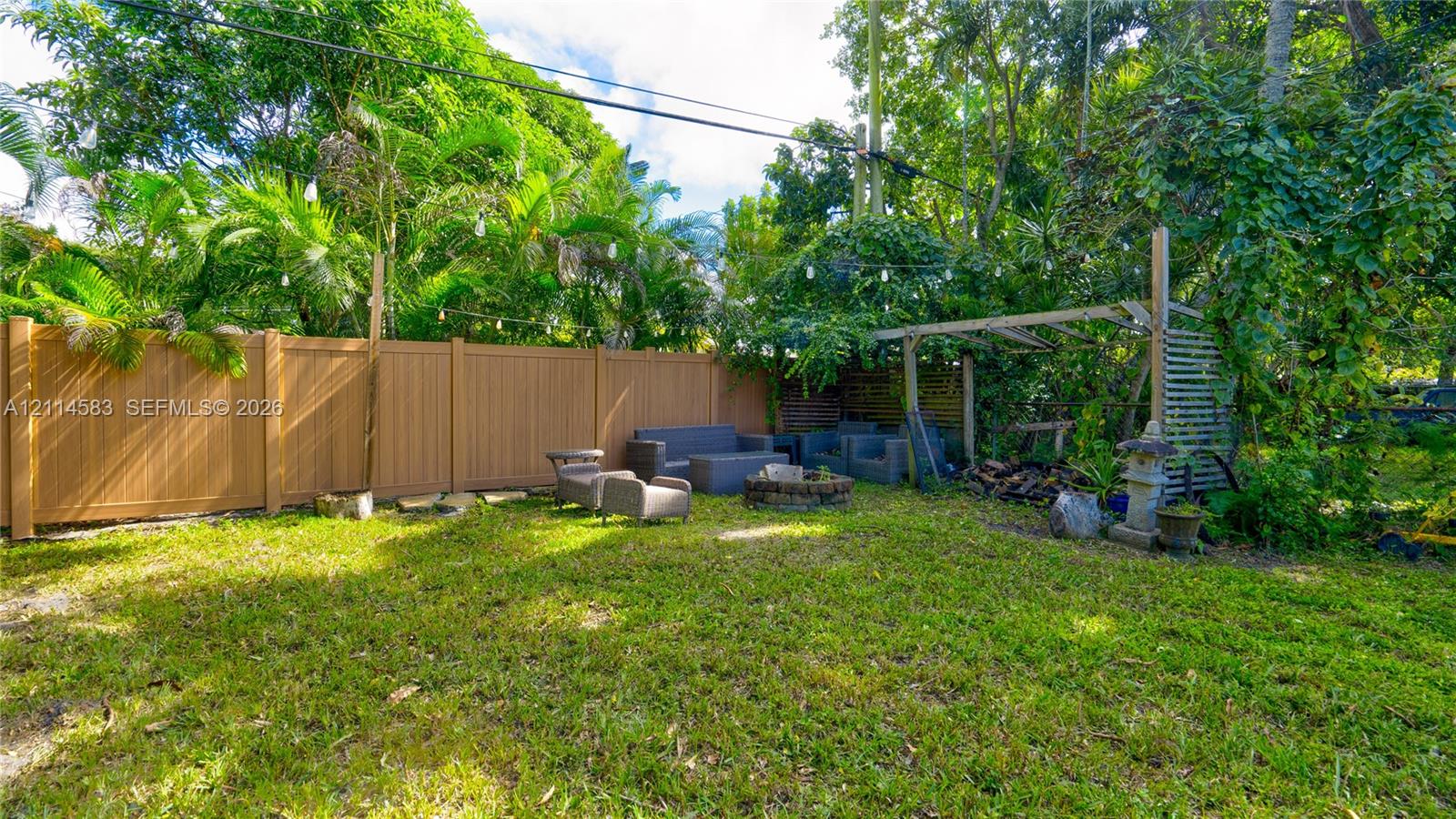 Backyard featuring a privacy fence, stone fire pit, and wooden pergola, surrounded by mature trees and lush greenery