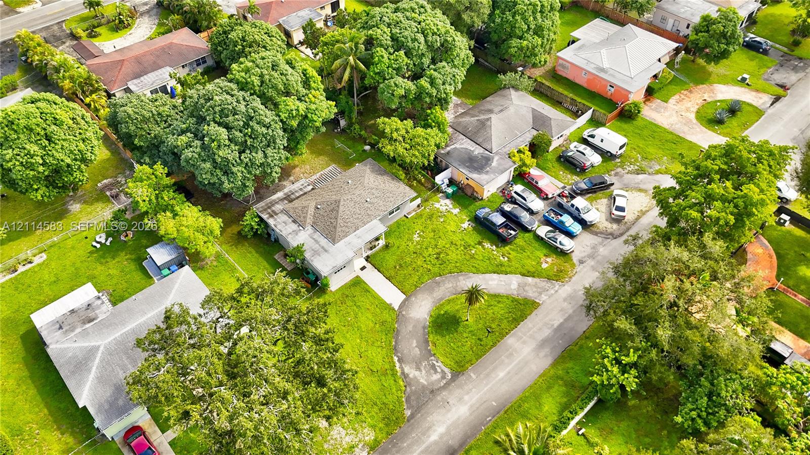 Aerial view of a residential lot featuring a hip roof structure, paved circular driveway, mature perimeter trees, and expansive lawn area