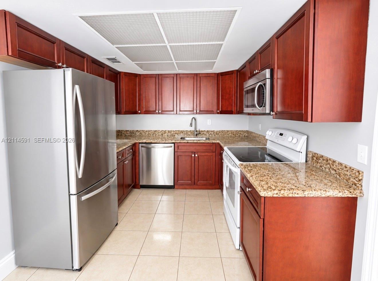 Kitchen featuring warm-tone cabinetry, speckled stone countertops, tile flooring, stainless steel appliances, and a recessed ceiling panel