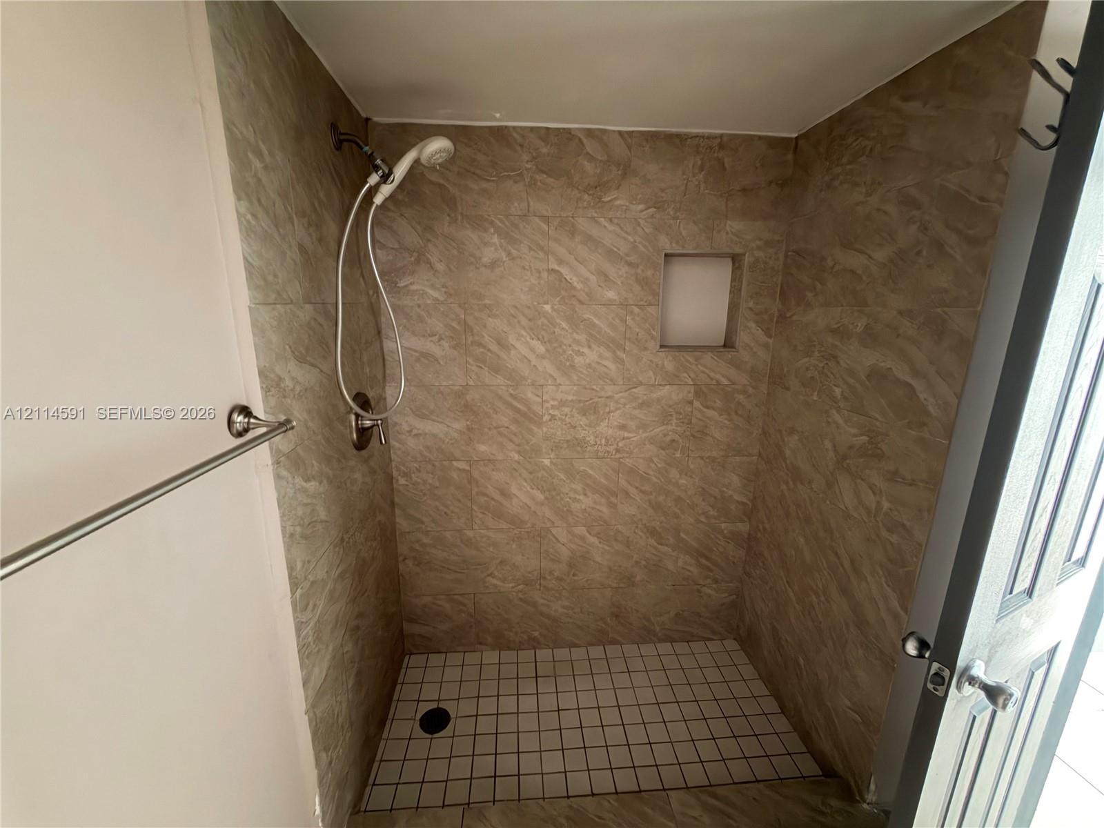 Walk-in shower featuring neutral stone-finish wall tile, a built-in recessed wall niche, a square-tile shower floor, and a polished chrome handheld shower fixture