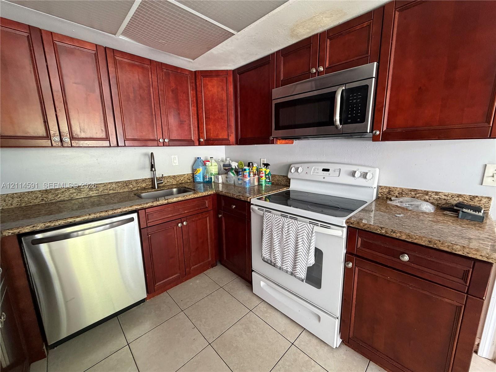 Kitchen featuring warm-tone wooden cabinetry, stone-finish countertops, stainless steel dishwasher, integrated over-the-range microwave, and tile flooring