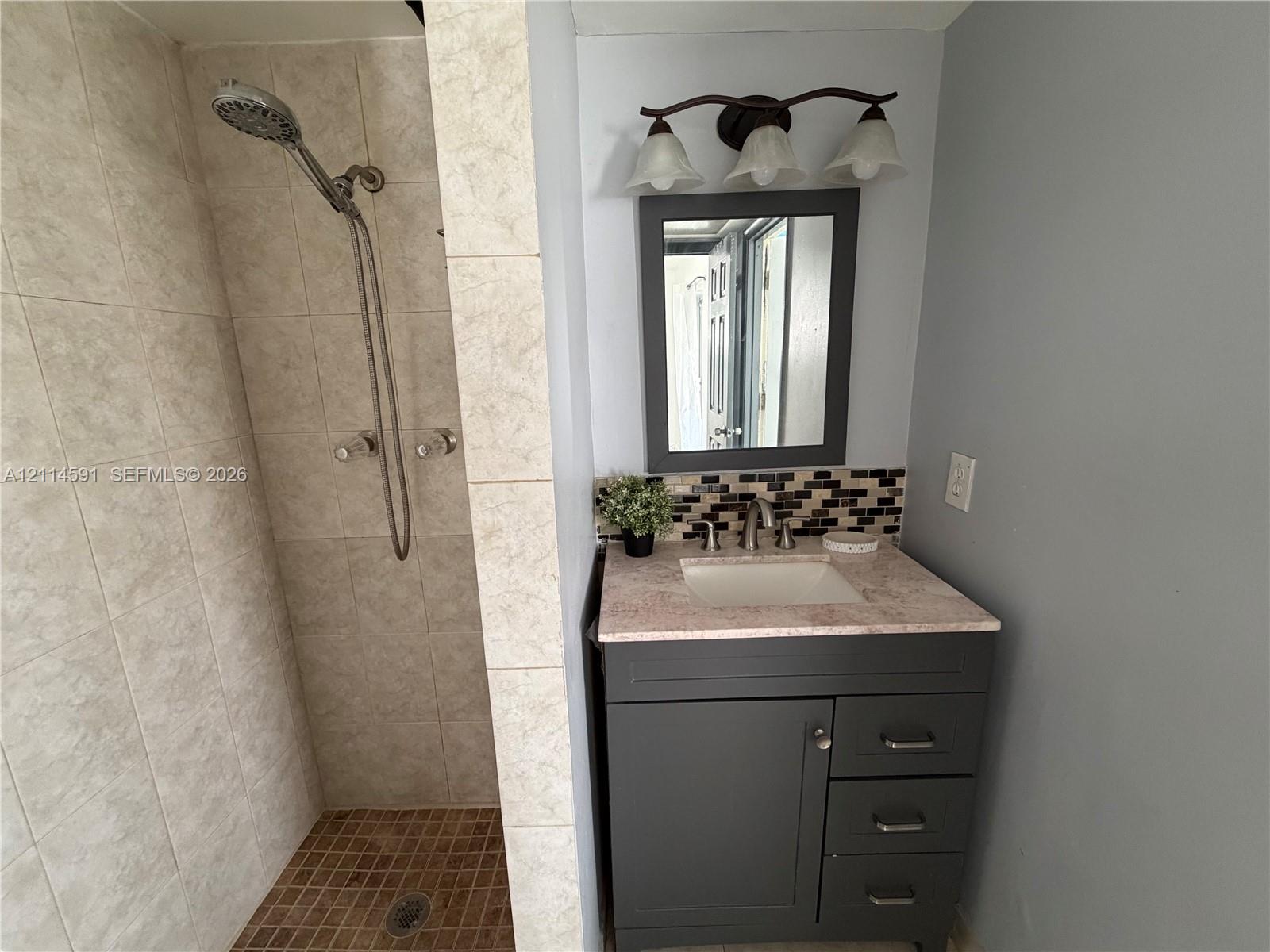Walk-in shower featuring tiled walls and a textured floor, vanity with a solid-surface countertop, rectangular undermount sink, multi-tone glass tile backsplash, and a three-light fixture