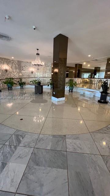 Grand foyer featuring polished stone flooring, custom-patterned wall finishes, a central structural column with a reflective finish, a multi-tiered crystal chandelier, and decorative metal railings Grand foyer featuring polished stone flooring, custom-patterned wall finishes, a central structural column with a reflective finish, a multi-tiered crystal chandelier, and decorative metal railings