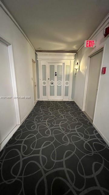 Hallway featuring patterned carpeting, paneled wood door with decorative molding, and wall-mounted sconce lighting Hallway featuring patterned carpeting, paneled wood door with decorative molding, and wall-mounted sconce lighting
