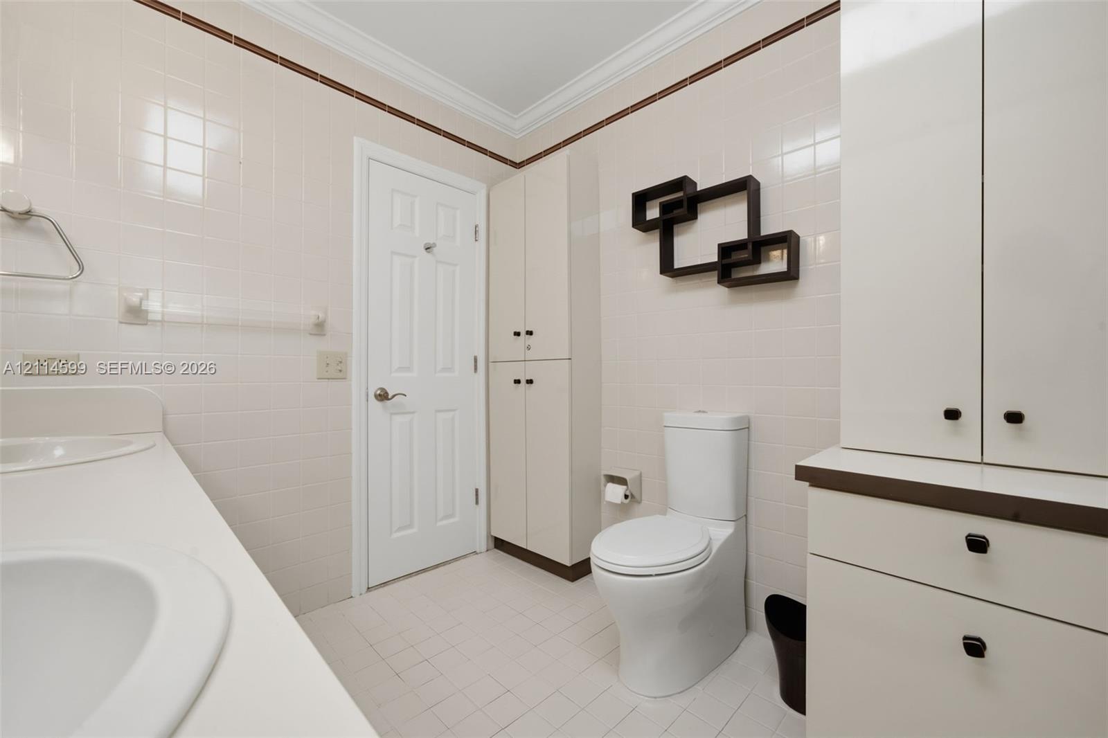 Bathroom featuring floor-to-ceiling ceramic wall tiling, dual-basin vanity with solid surface countertop, crown molding, and extensive built-in cabinetry