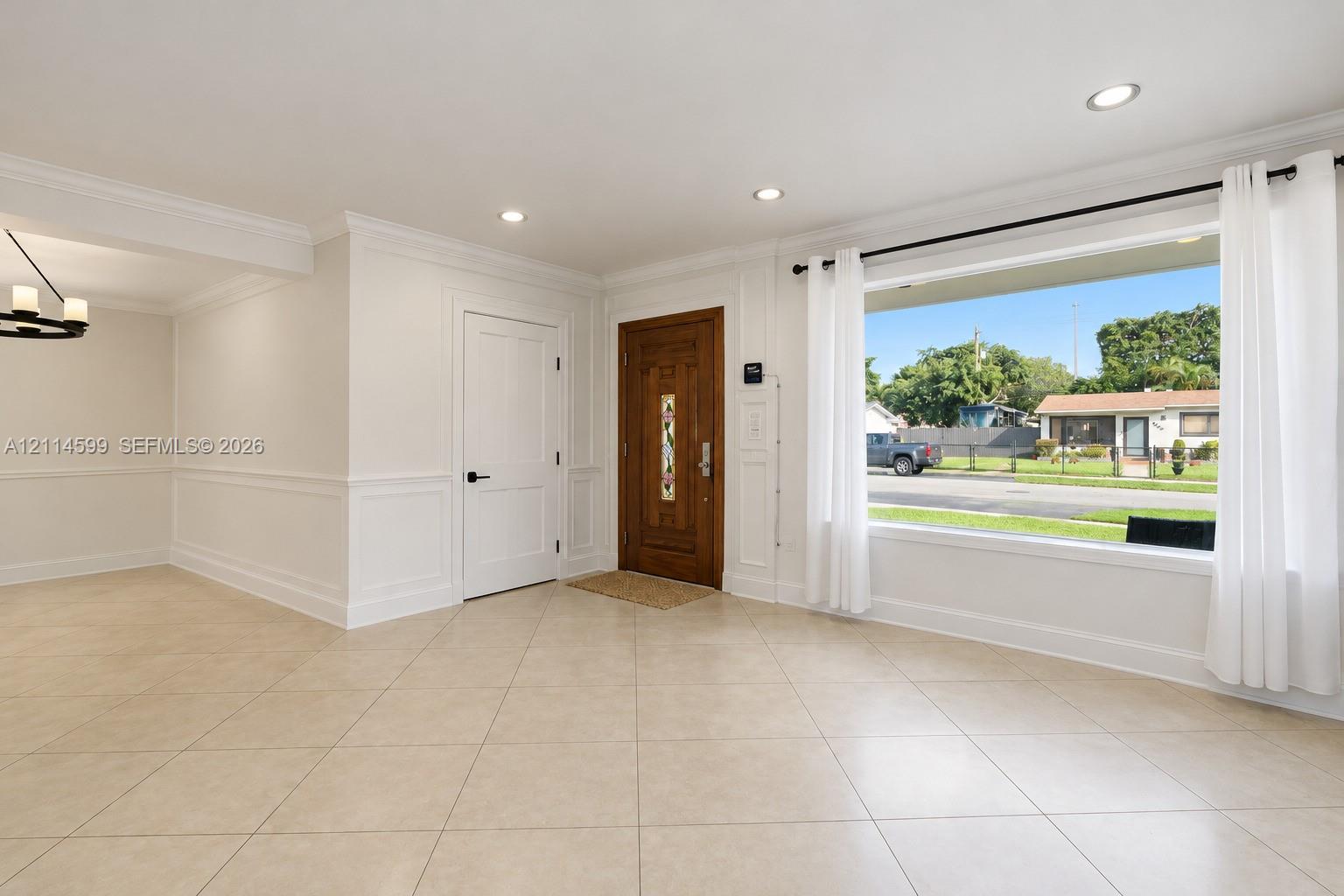 Neutral tile flooring, expansive picture window, wainscoting detail, recessed lighting, and solid wood-finish entry door with decorative glass inlay