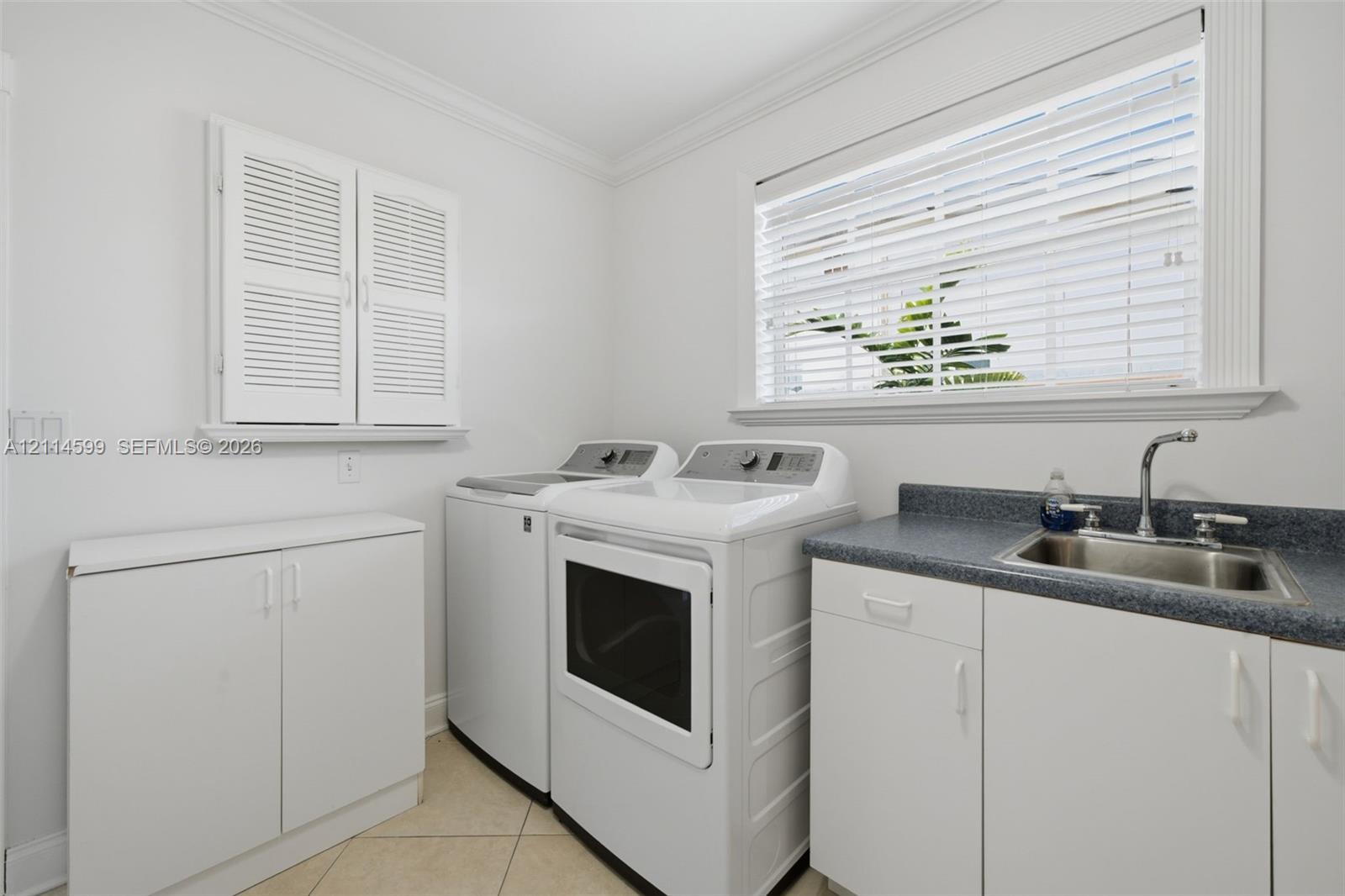 Laundry room featuring custom white cabinetry, stone-finish countertops, an integrated stainless steel sink, tile flooring, and louvered shutters