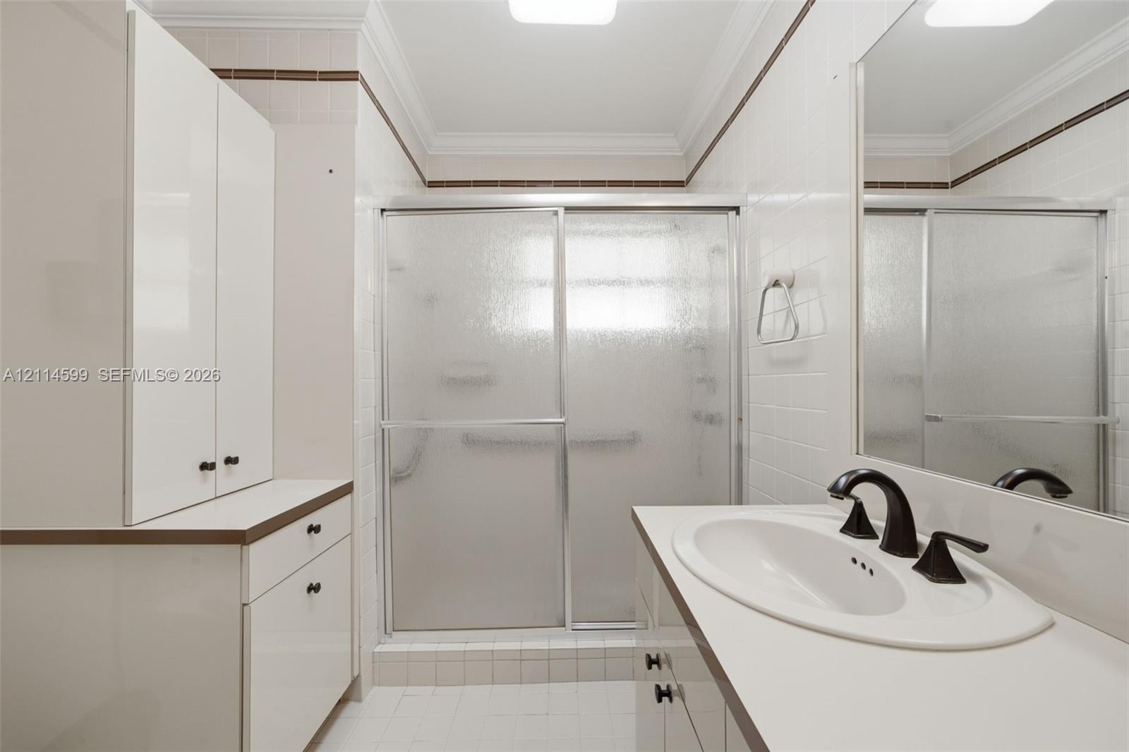 Bathroom featuring white cabinetry, solid-surface countertop, integrated oval sink with dark-finish hardware, glass-enclosed shower, and crown molding