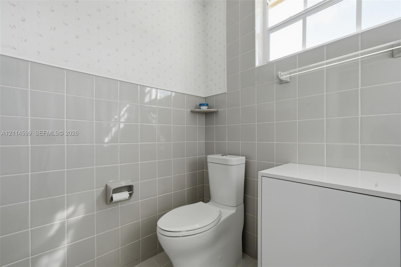 Glossy gray wall tile, dual-flush toilet, white cabinet with solid-surface top, integrated corner shelving, and multi-pane window