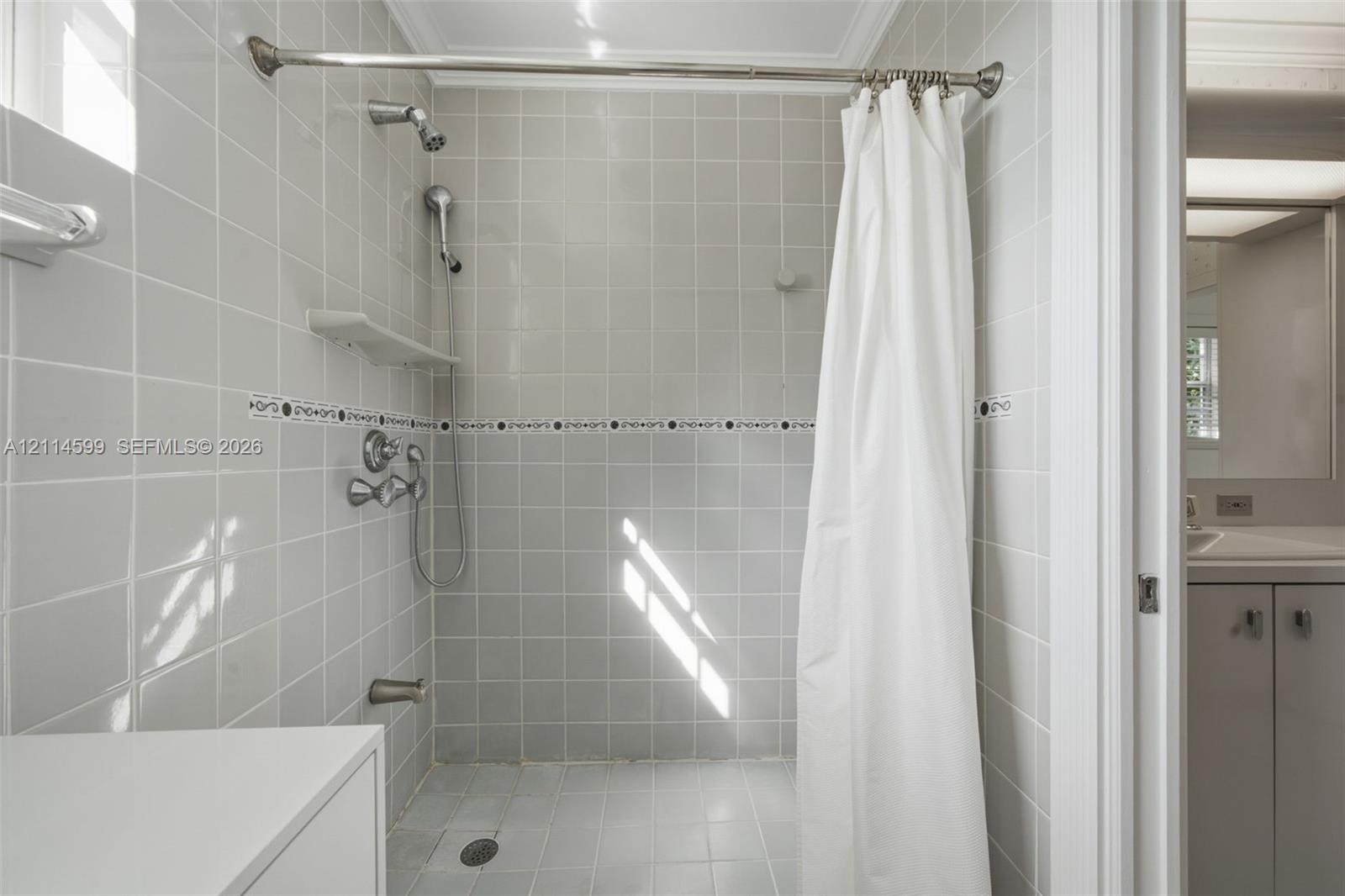 Shower enclosure featuring white square tile wall surrounds with a decorative accent border, chrome finish wall-mounted fixtures, and a built-in corner shelf